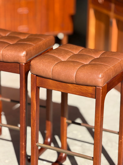 Three Mid Century Australian 1960s Barstools by Gerald Easden, bar height variant, feature square seats with brown tufted leather and sturdy wooden frames, capturing classic style with warm wood accents in bright sunlight.