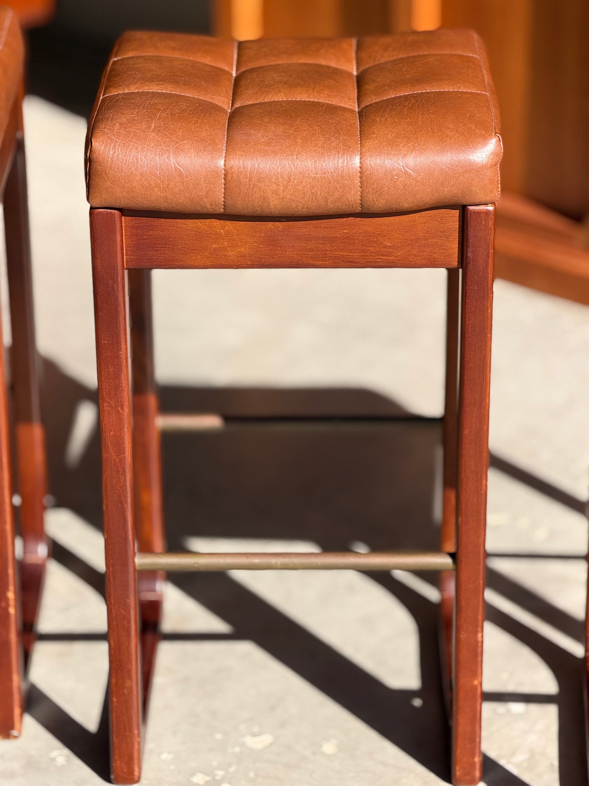 A set of three Mid Century Australian 1960s barstools by Gerald Easden (Revive, Bar Height) with square, brown leather stitched seats and wooden frames, inspired by mid-century design.