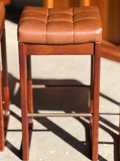 A set of three Mid Century Australian 1960s barstools by Gerald Easden (Revive, Bar Height) with square, brown leather stitched seats and wooden frames, inspired by mid-century design.