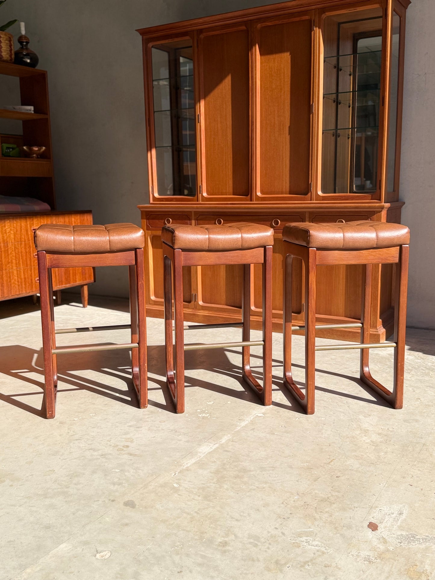A set of three Mid Century Australian 1960s Barstools by Gerald Easden (bar height, Revive) with brown leather seats stand in a row on sunlit concrete, with wooden cabinets and a glass hutch evoking mid-century style in the background.