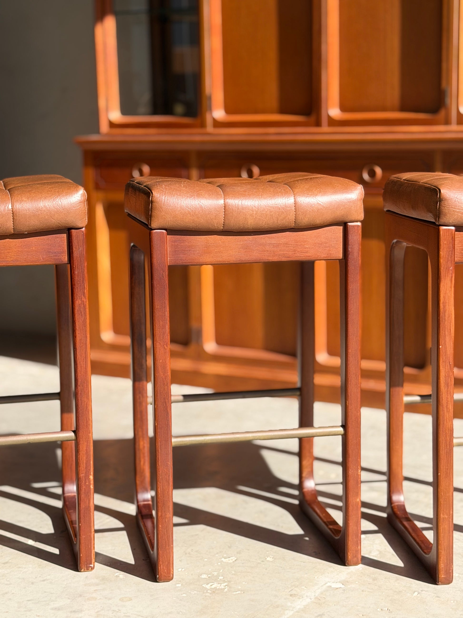 Three Mid Century Australian 1960s Barstools by Gerald Easden (bar height, set of 3, Revive) with brown leather seats are arranged in sunlight, their shadows on the floor. A matching wooden cabinet echoes the vintage style in the background.