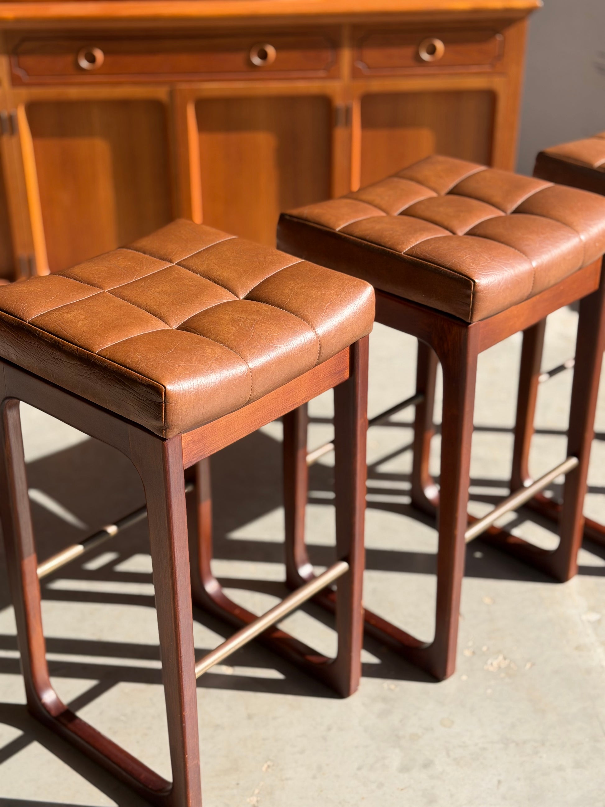 Three Mid Century Australian 1960s Barstools by Gerald Easden (Bar Height, Set of 3) are lined up, their brown tufted leather seats and wooden frames casting shadows on the concrete floor near a matching mid-century style cabinet.