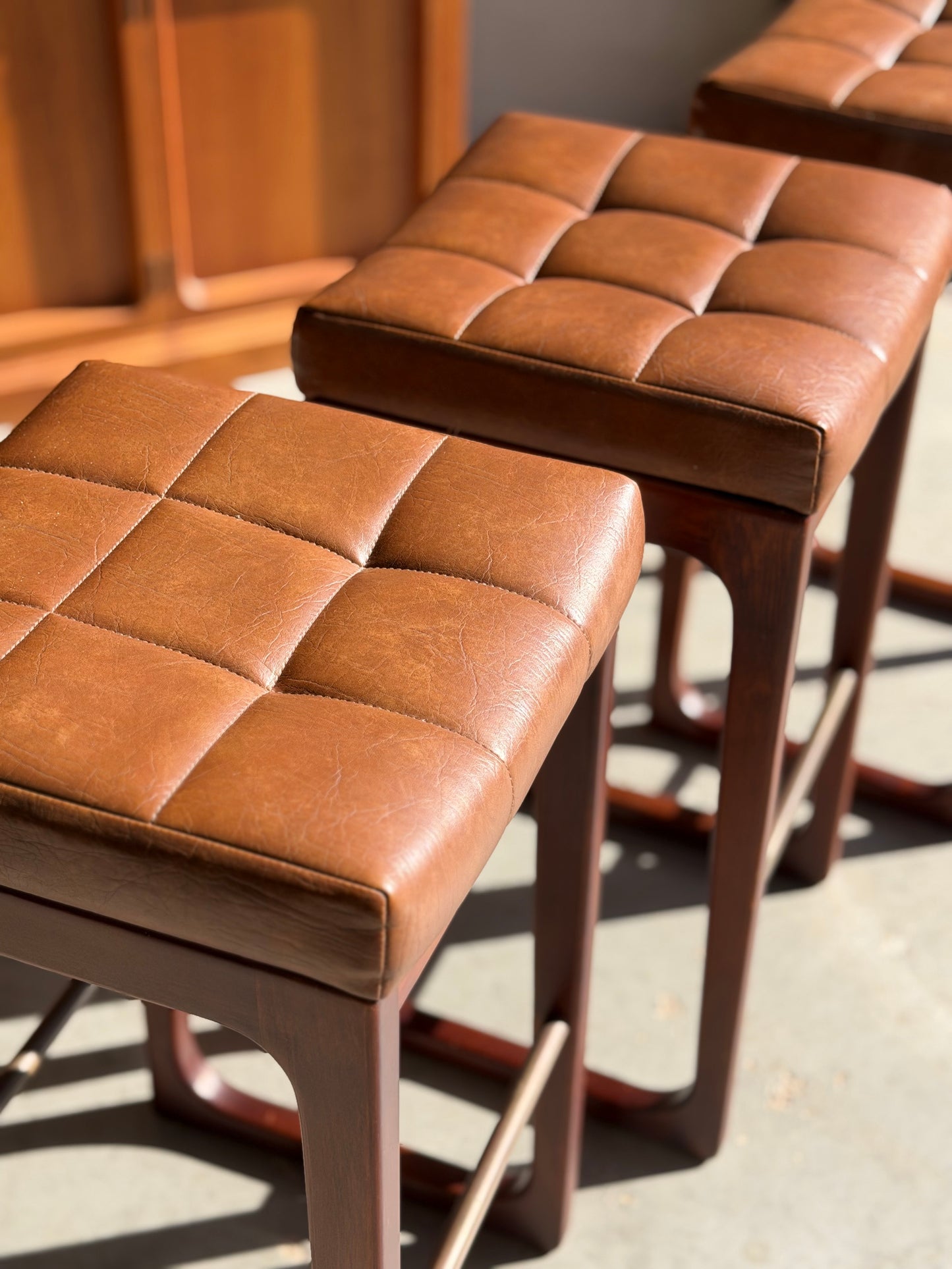 A set of three Mid Century Australian 1960s Barstools by Gerald Easden (Bar Height Variant, Revive) featuring brown tufted leather seats and wooden legs are arranged in sunlight, highlighting their texture in classic mid-century style.