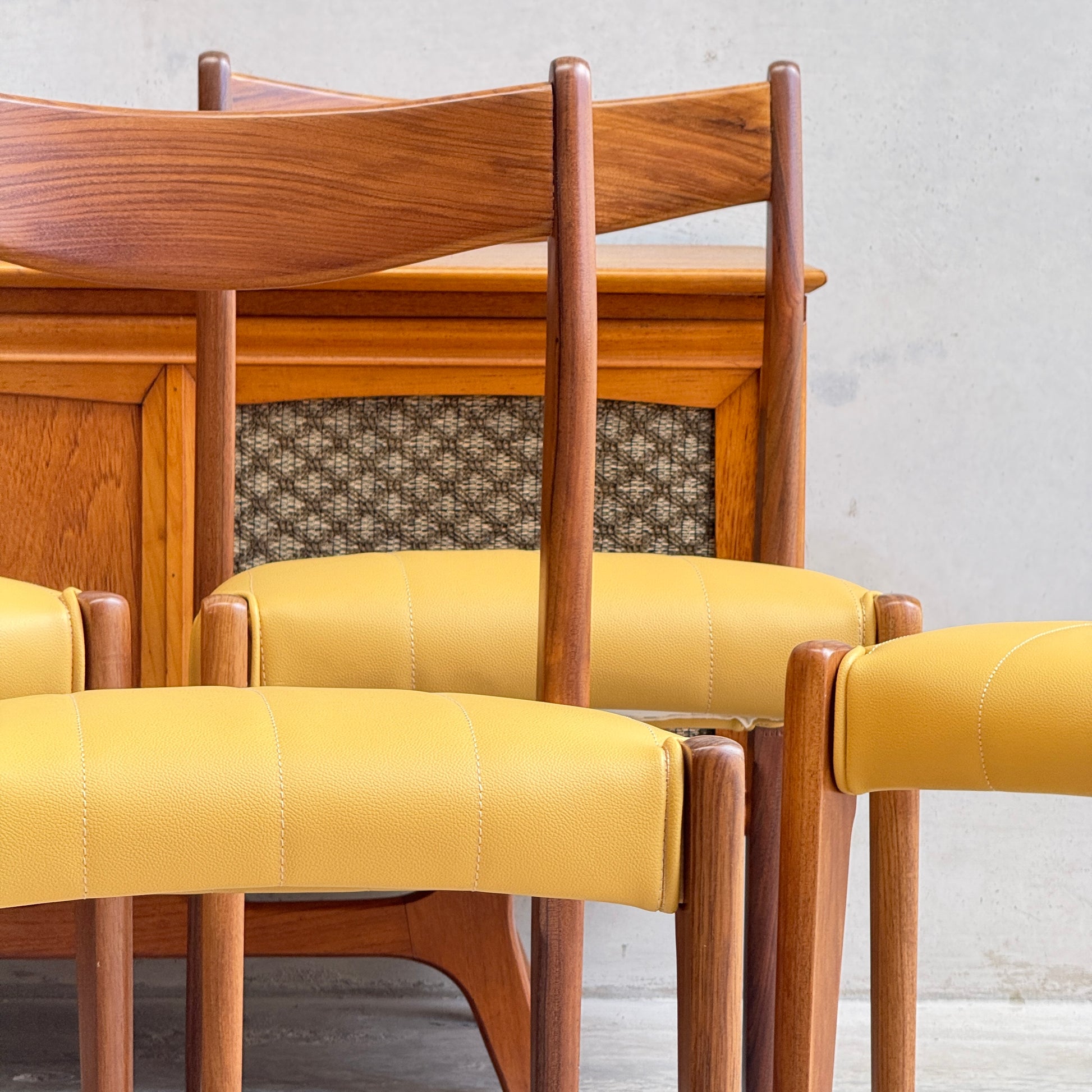 A set of four fully restored THB 'Stockholm' dining chairs with yellow seats and curved wooden backs is arranged in front of a wooden sideboard with a woven panel, all set against a light gray wall.
