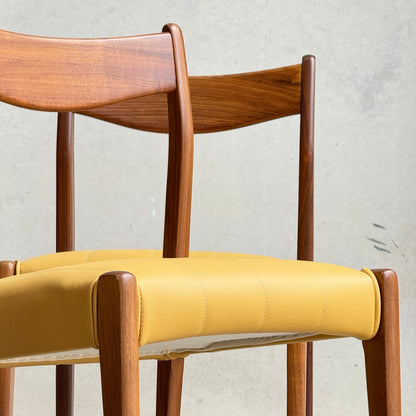 A set of four fully restored THB 'Stockholm' dining chairs (Rediscover) with curved backrests and mustard yellow cushions are stacked against a light gray wall.