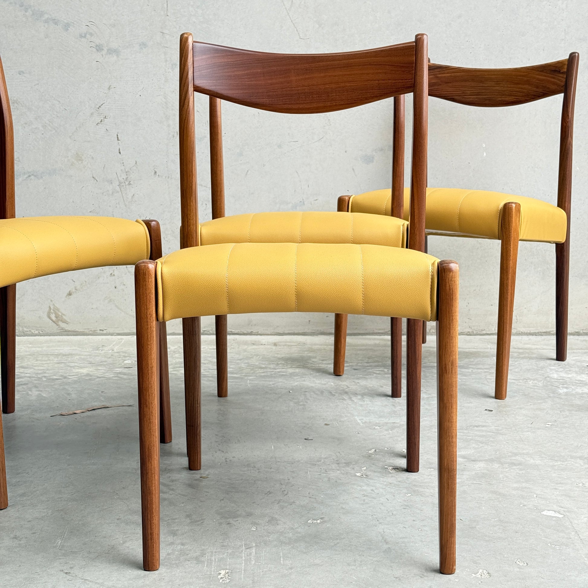 Four fully restored THB 'Stockholm' dining chairs are arranged on a concrete floor against a plain, light gray wall, showcasing their mid-century modern design and curved backrests.