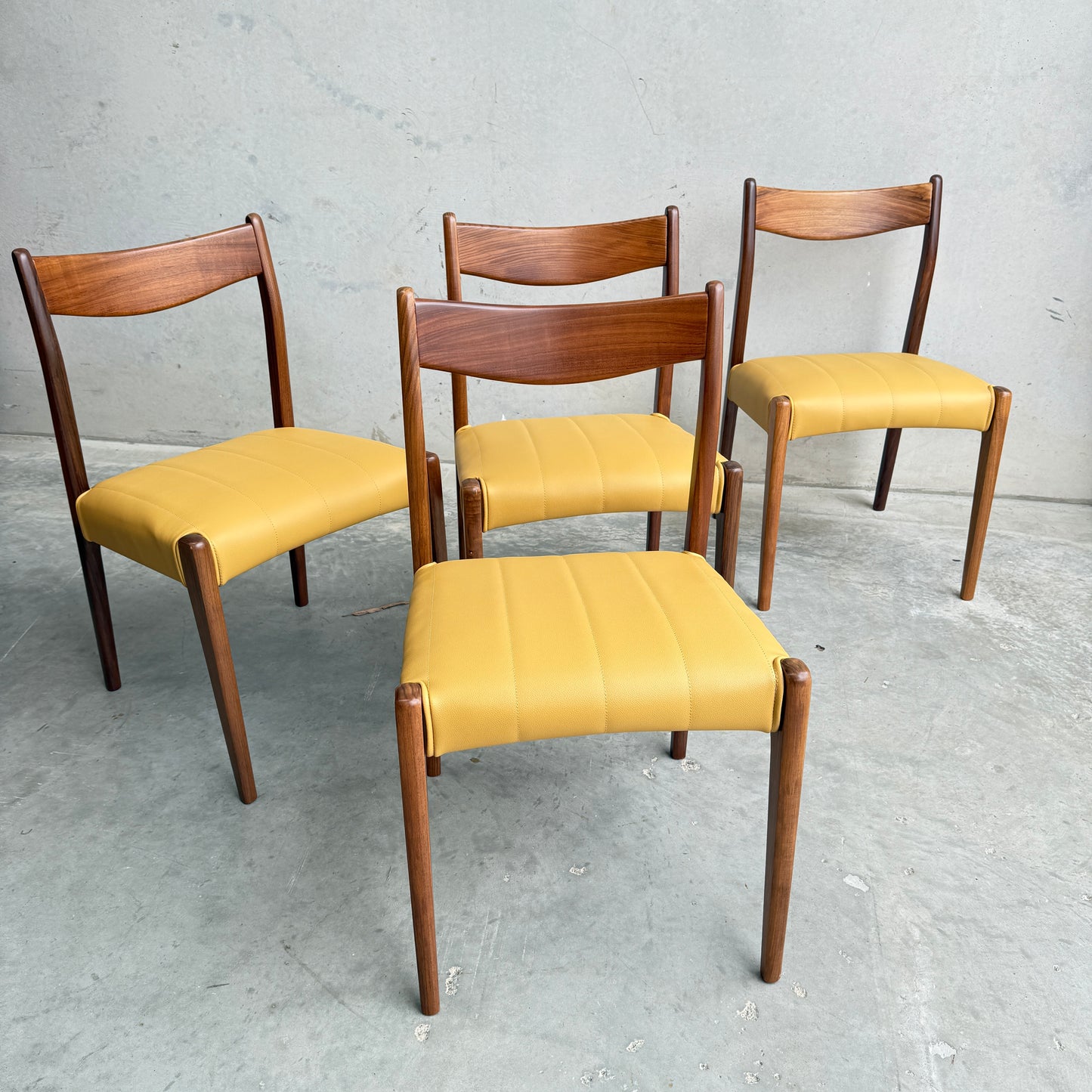 A set of four fully restored THB 'Stockholm' dining chairs (Rediscover) with curved wooden backrests and light yellow upholstered seats are arranged on a gray concrete floor against a plain light gray wall.