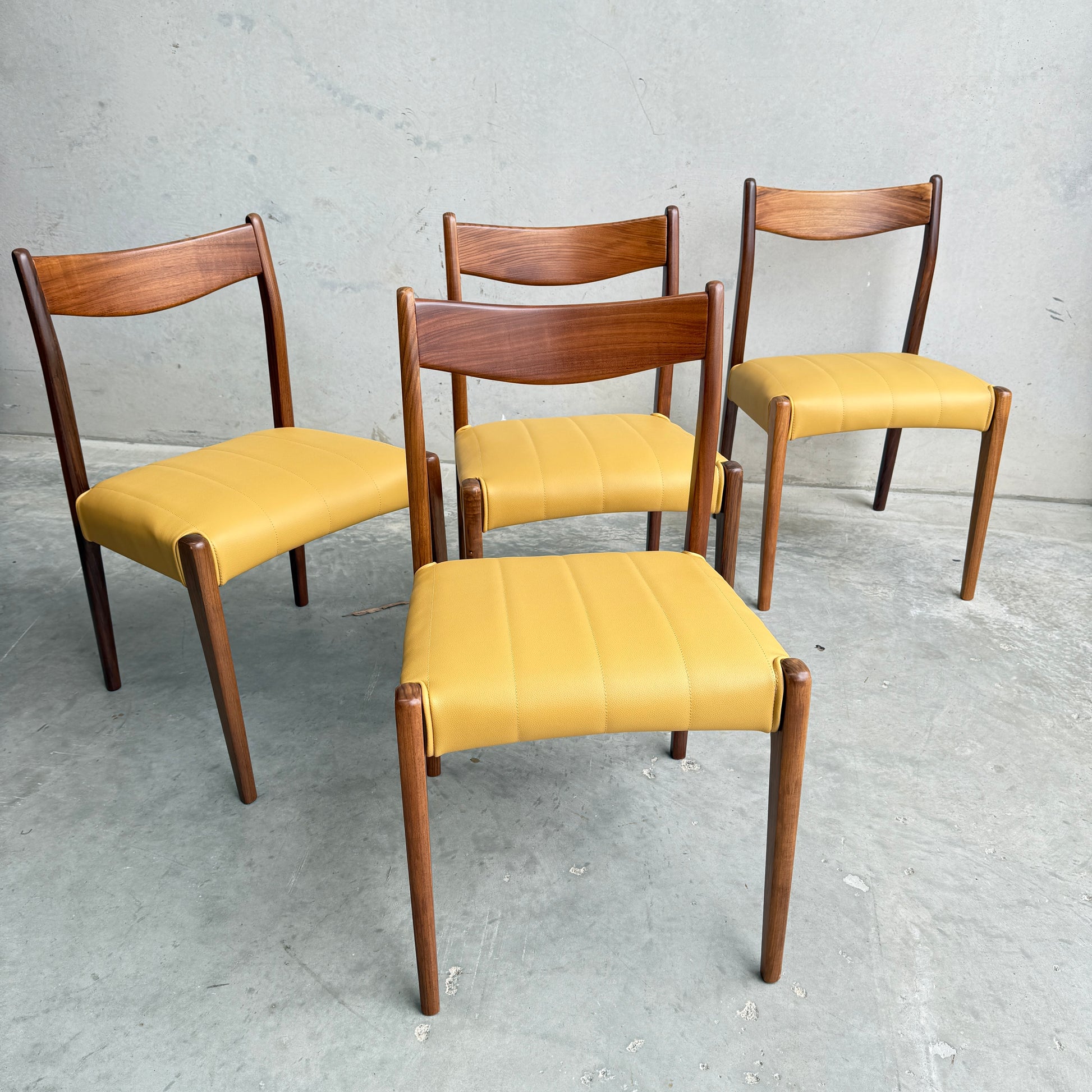 A set of four fully restored THB 'Stockholm' dining chairs (Rediscover) with curved wooden backrests and light yellow upholstered seats are arranged on a gray concrete floor against a plain light gray wall.