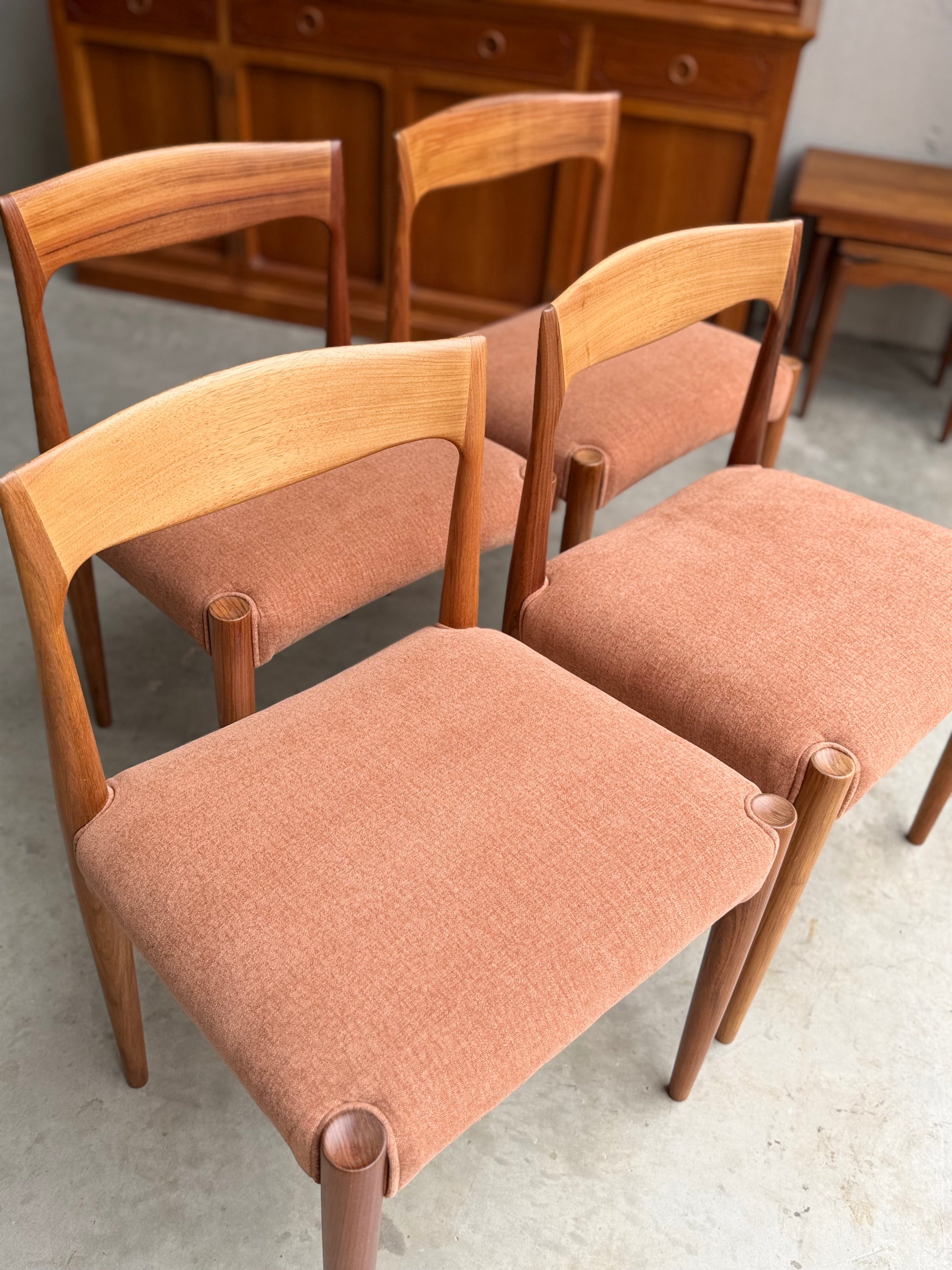 A set of four fully restored THB 'Milan' dining chairs (Rediscover) with curved backs and light brown upholstered seats are arranged on a light floor, with a wooden sideboard and small table in the background.