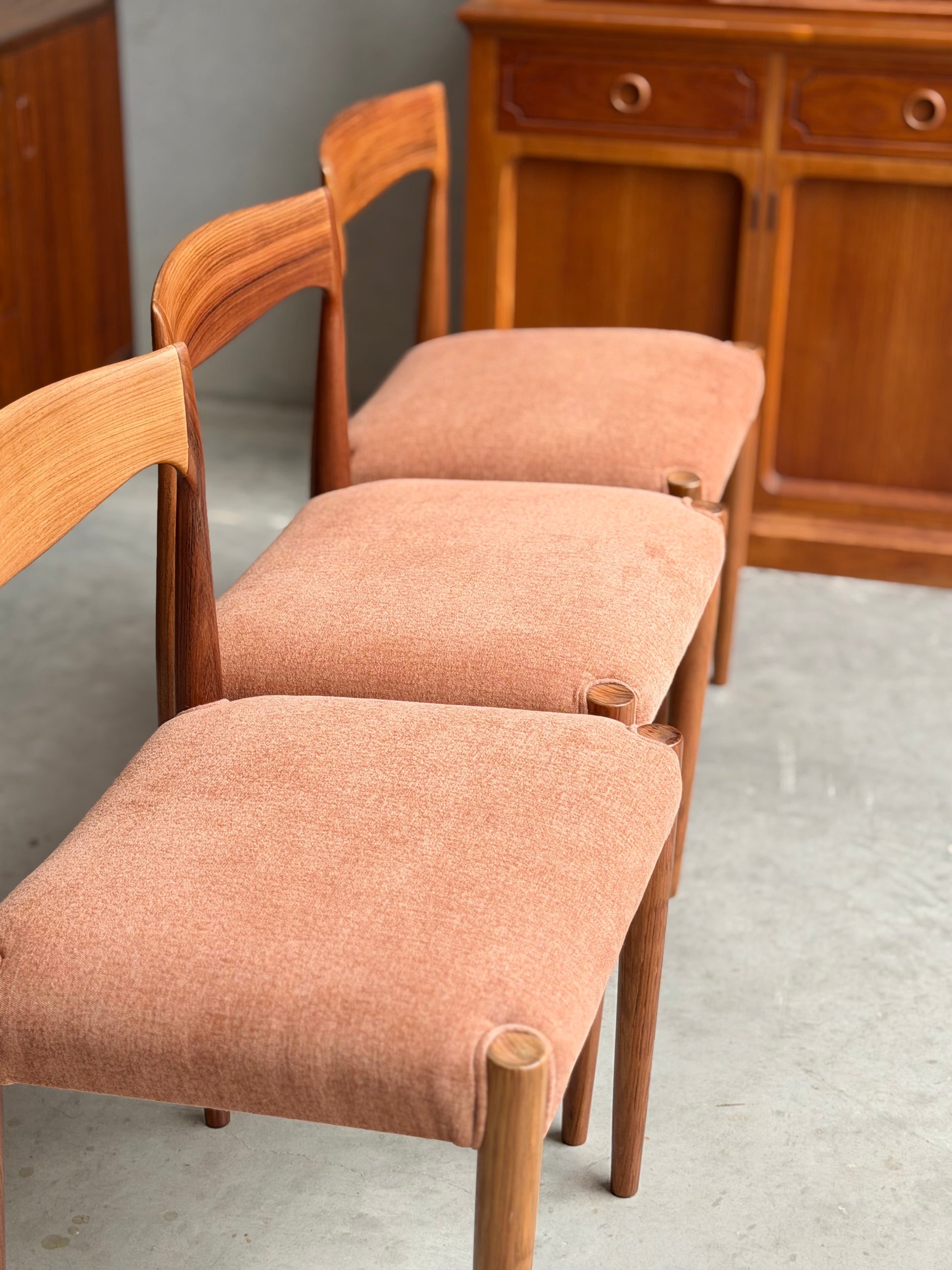 Four fully restored THB 'Milan' dining chairs from Rediscover, featuring light brown upholstered seats, are arranged on a light gray concrete floor with a wooden cabinet with drawers and doors in the background.