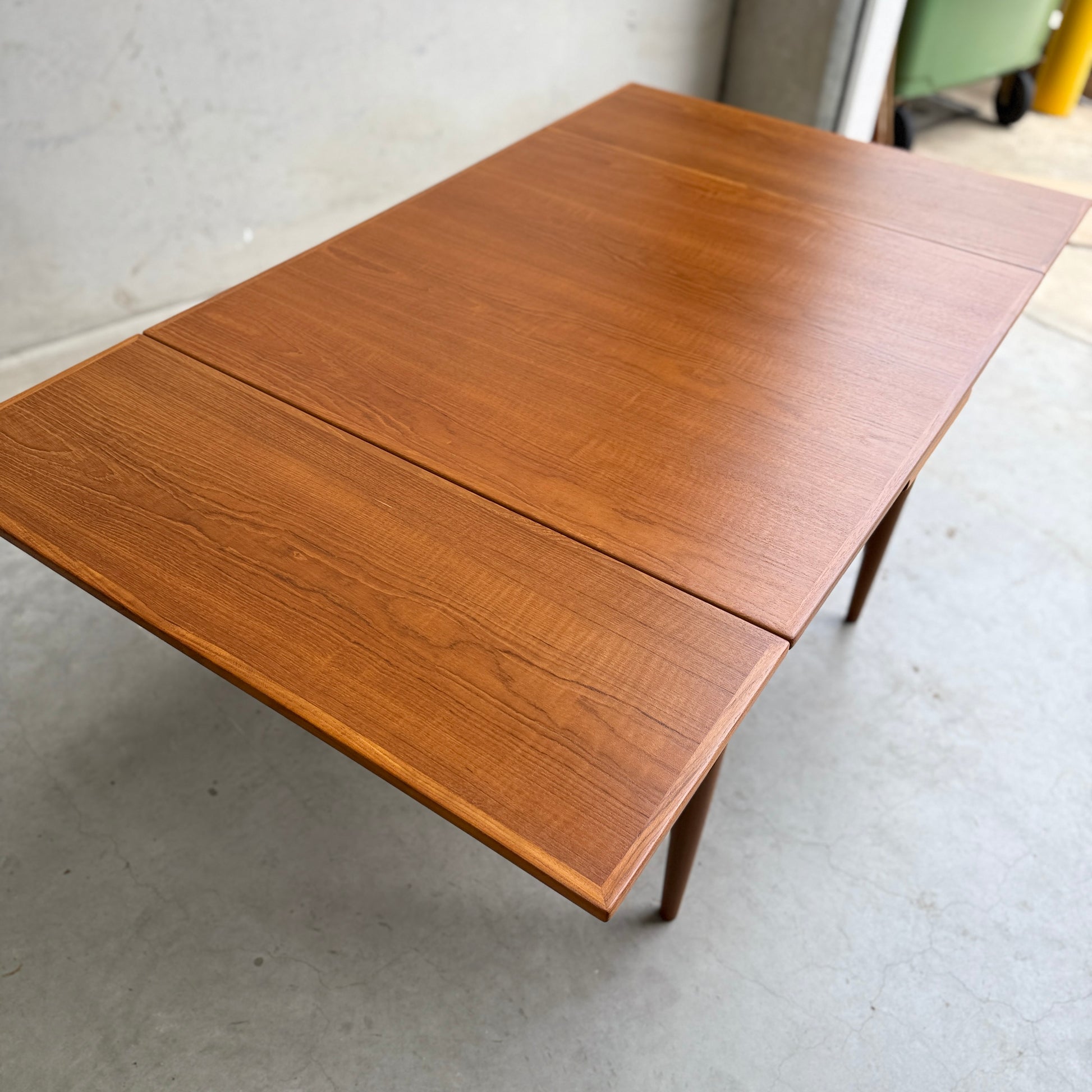 The Fully Restored Parker 140 Square Extension Table (Rediscover) features a smooth teak finish and visible wood grain, standing against a plain indoor setting with a concrete floor to highlight its timeless mid-century modern style.