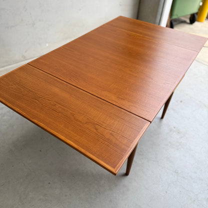 The Fully Restored Parker 140 Square Extension Table (Rediscover) features a smooth teak finish and visible wood grain, standing against a plain indoor setting with a concrete floor to highlight its timeless mid-century modern style.