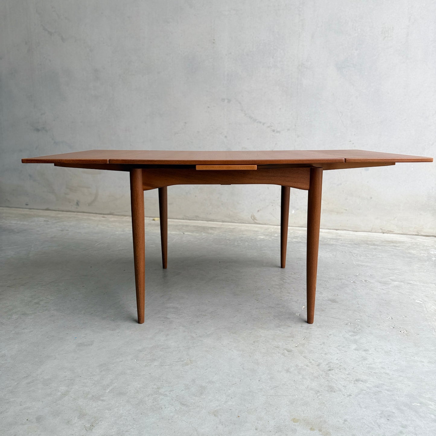 The Fully Restored Parker 140 Square Extension Table (Rediscover) features angled legs and a central drawer, set against a light gray wall and concrete floor to highlight its clean, minimalist mid-century modern design.