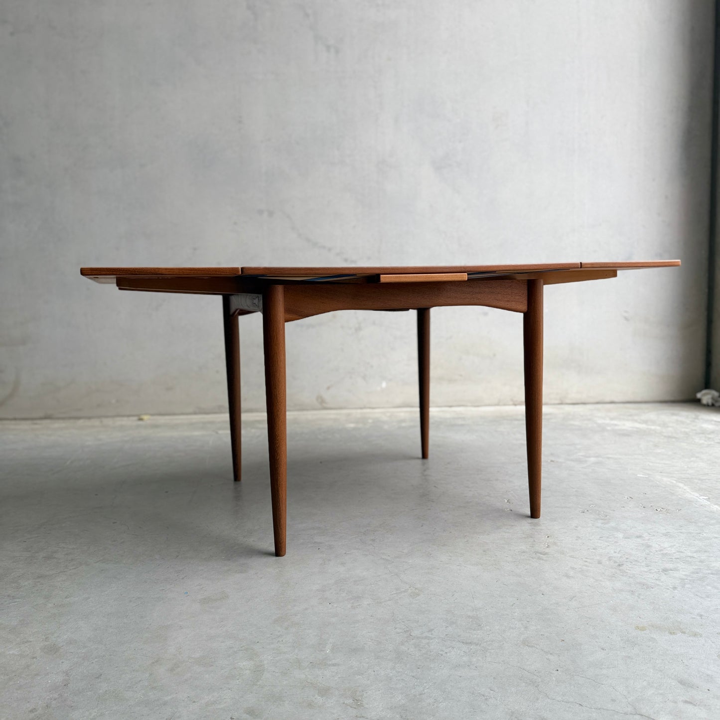 A Fully Restored Parker 140 Square Extension Table (Rediscover) with tapered legs and drop-leaf sides stands on a smooth concrete floor against a plain gray wall.