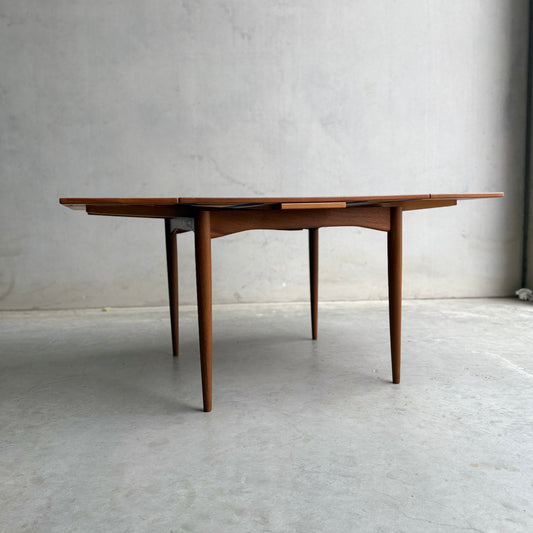 A Fully Restored Parker 140 Square Extension Table (Rediscover) with tapered legs and drop-leaf sides stands on a smooth concrete floor against a plain gray wall.