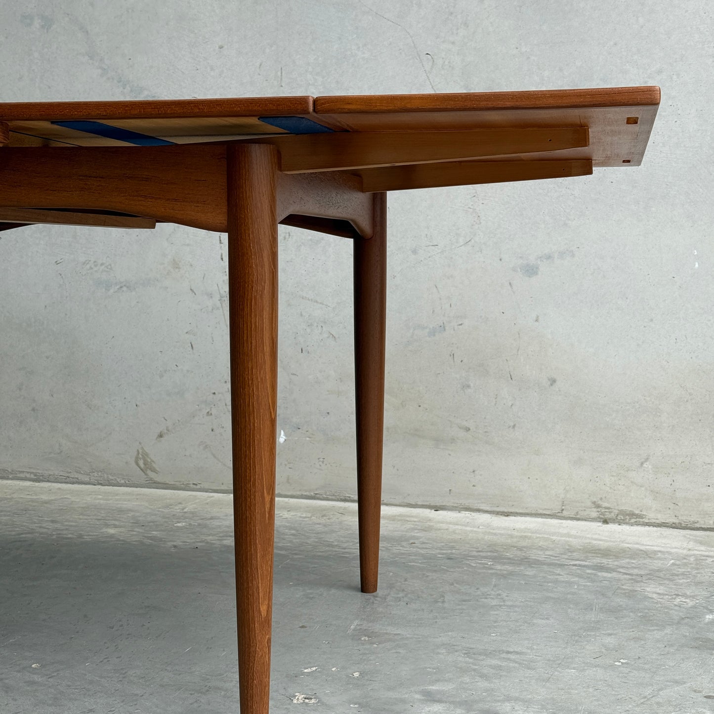 A close-up of the Fully Restored Parker 140 Square Extension Table (Rediscover) with tapered legs and an extending leaf, set against a plain light gray concrete wall and floor.