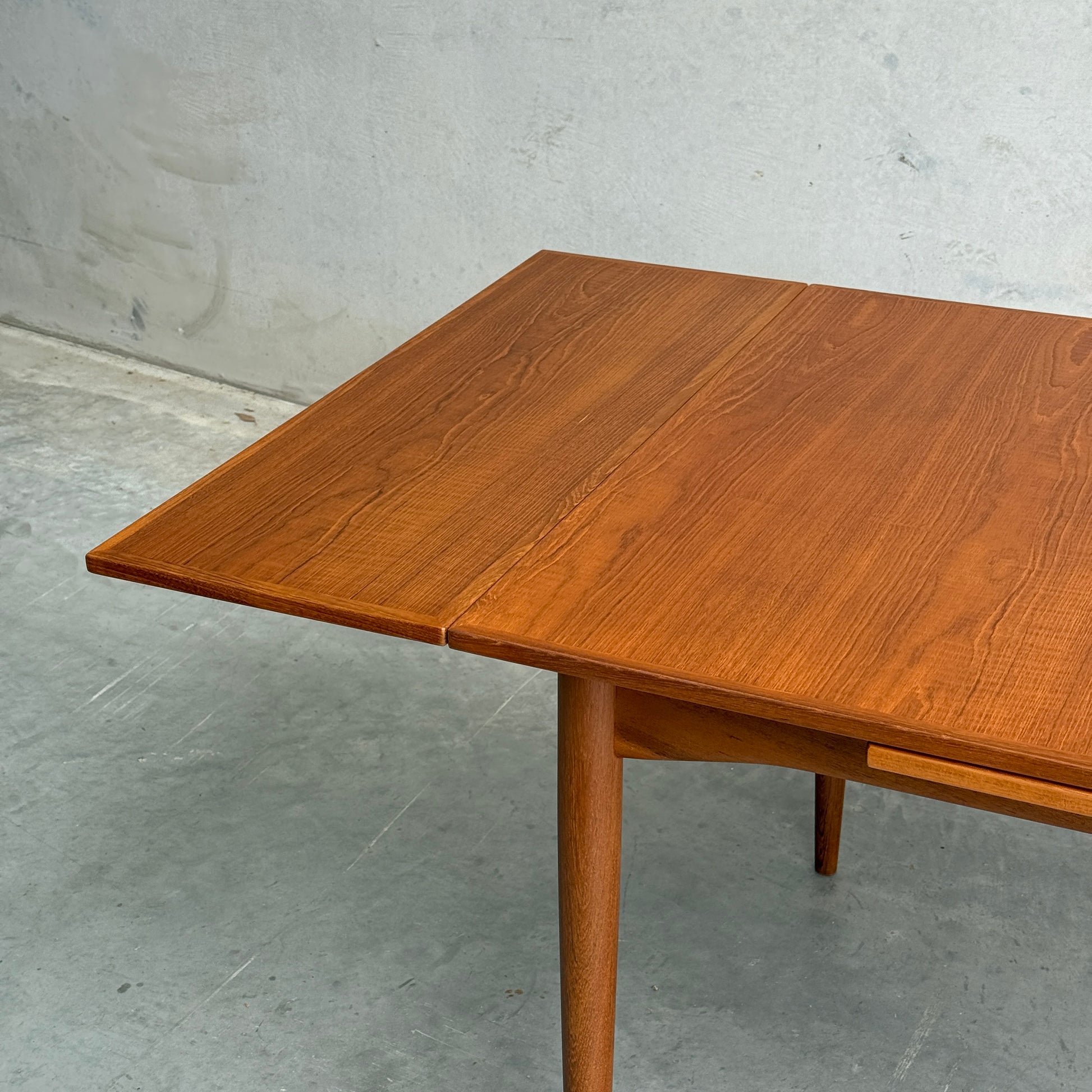 A close-up of the Fully Restored Parker 140 Square Extension Table (Rediscover) showcases its rich brown teak finish, clean lines, and visible wood grain, standing on tapered legs against a light gray wall and concrete floor.