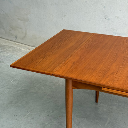 A close-up of the Fully Restored Parker 140 Square Extension Table (Rediscover) showcases its rich brown teak finish, clean lines, and visible wood grain, standing on tapered legs against a light gray wall and concrete floor.