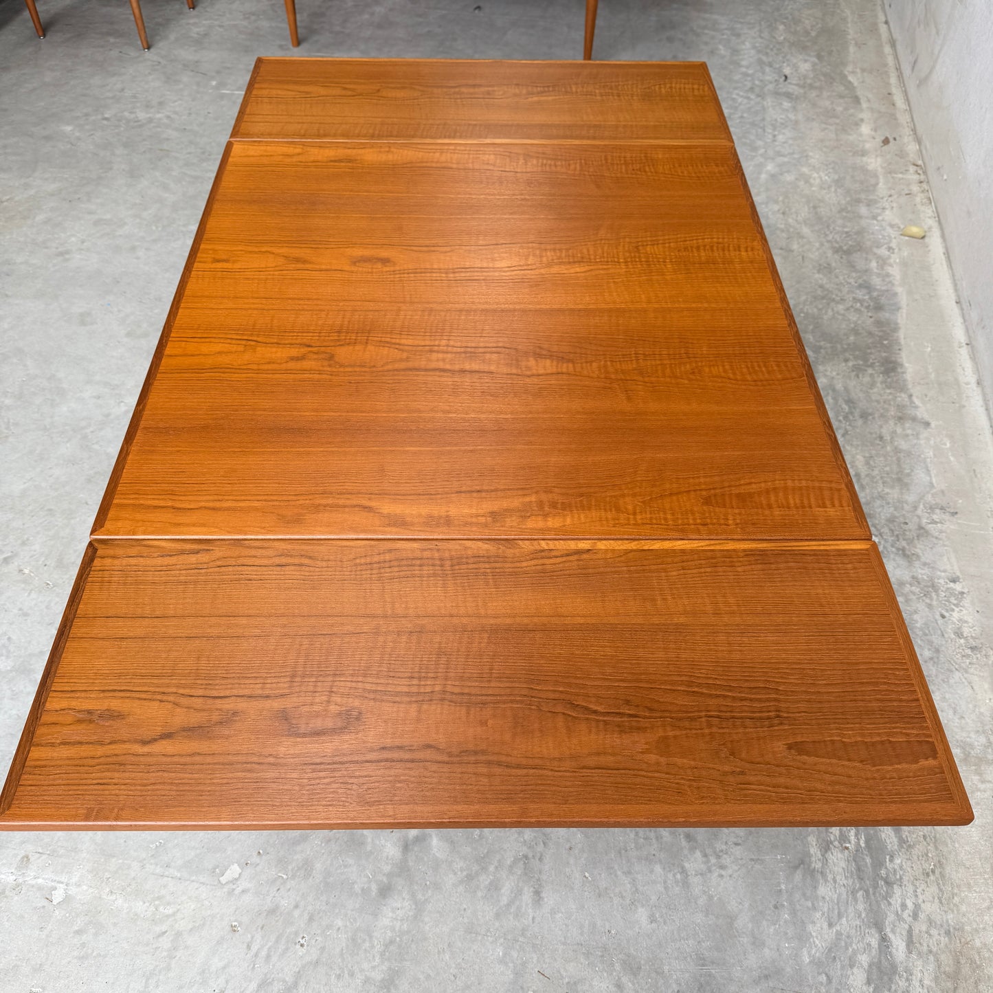 A Fully Restored Parker 140 Square Extension Table (Rediscover), featuring a smooth, polished teak surface and visible wood grain, sits on a gray concrete floor in a minimally furnished room.