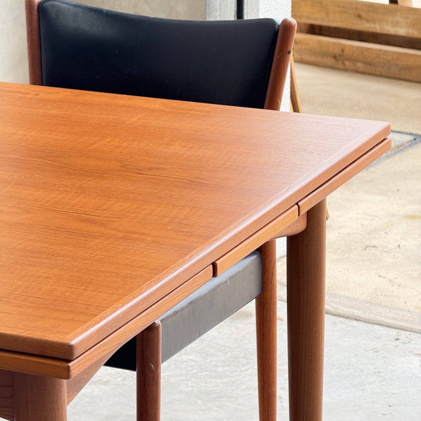 Close-up of the Fully Restored Parker 140 Square Extension Table (Rediscover) with rounded edges and a smooth finish, set indoors near a concrete floor, in front of a black cushioned chair by a window or doorway.