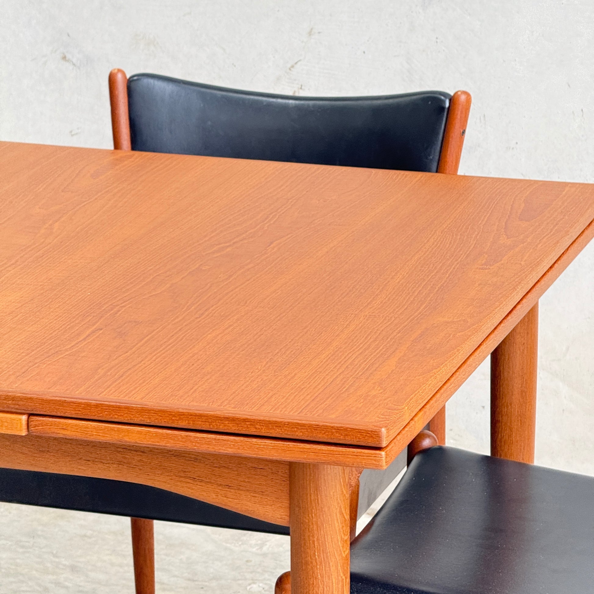 A close-up of the Fully Restored Parker 140 Square Extension Table (Rediscover) with rounded legs and a smooth surface, paired with two black curved-back chairs, set against a plain light-colored wall.