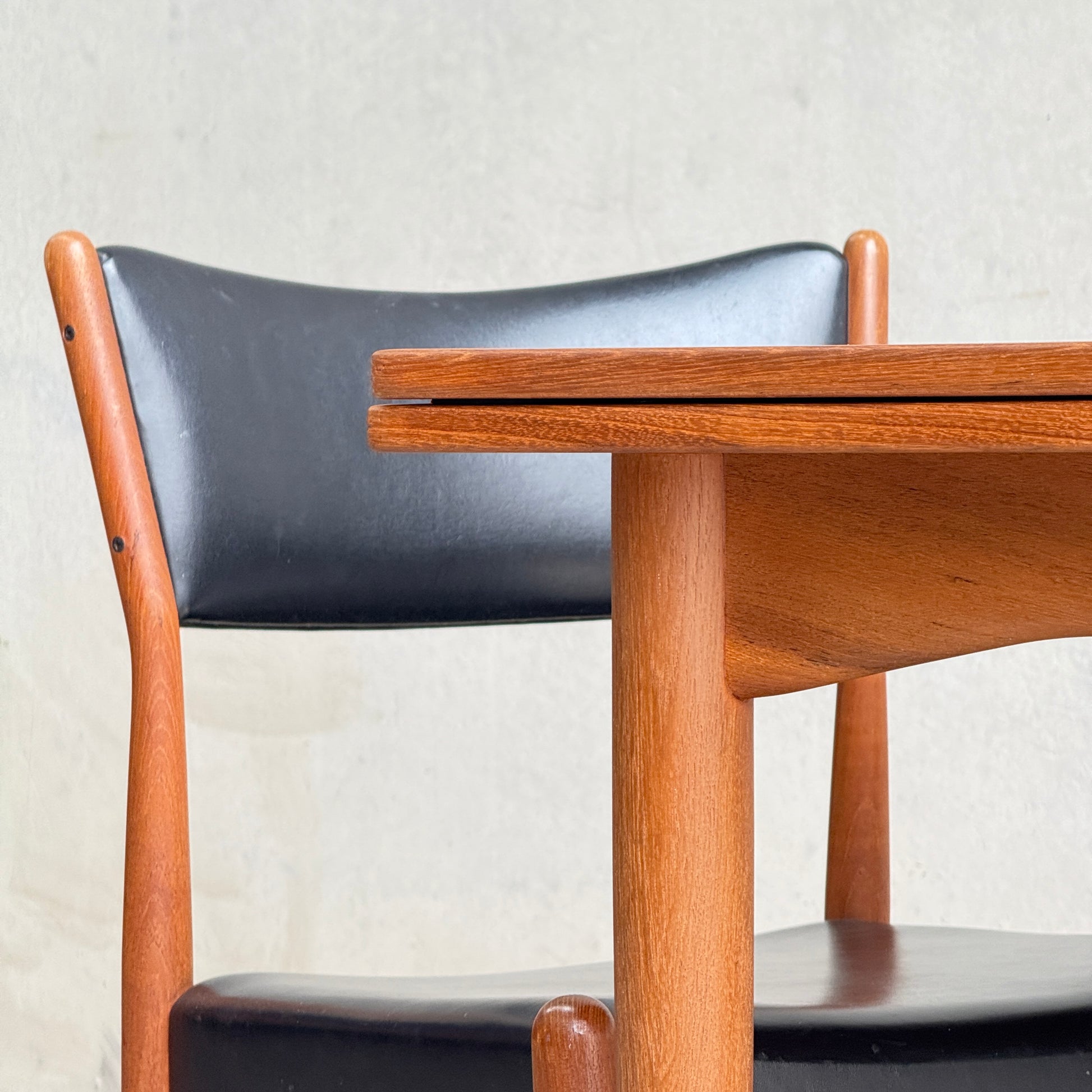 A close-up of a mid-century modern wooden chair with a black seat and backrest, placed behind the Fully Restored Parker 140 Square Extension Table (Rediscover) against a plain light-colored wall.