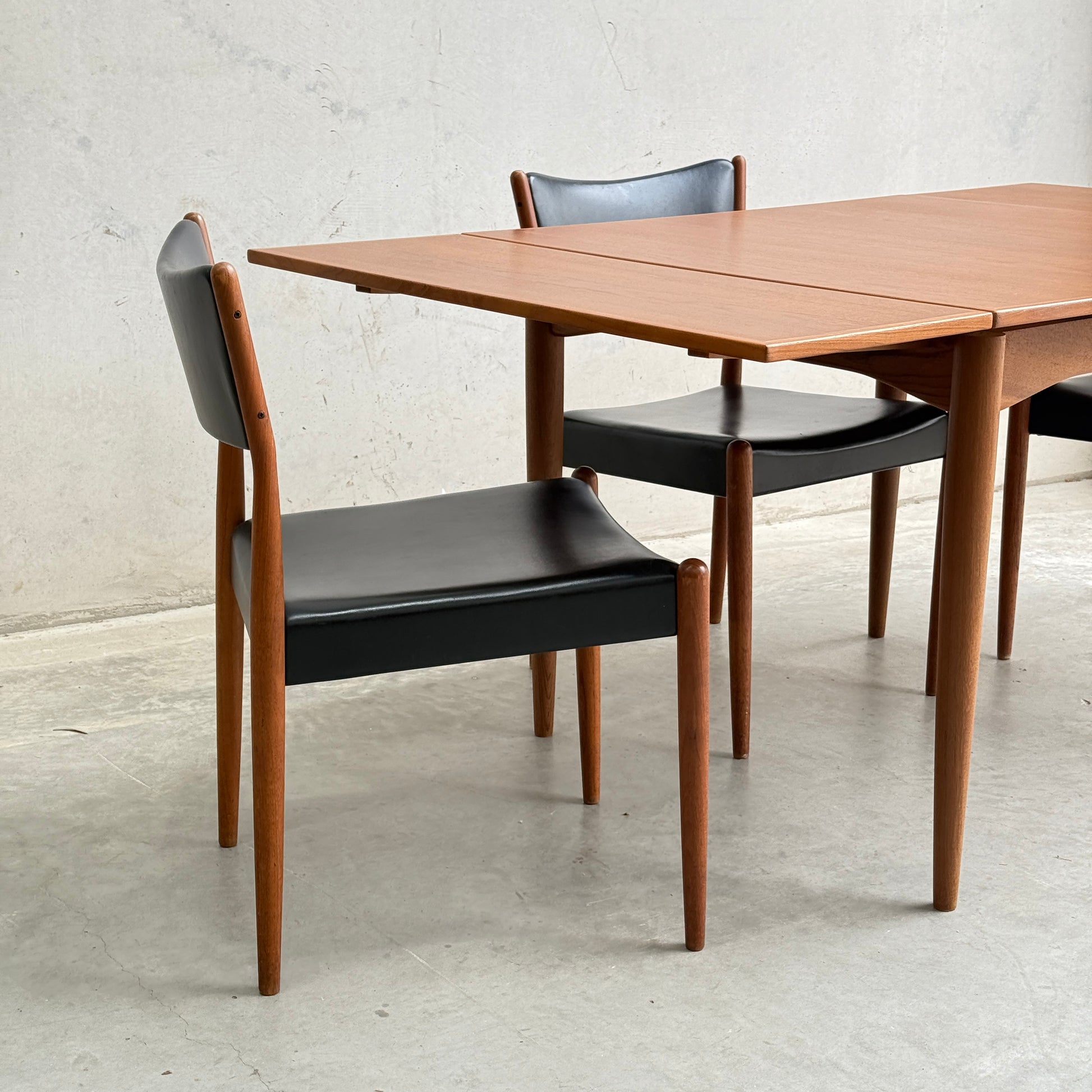 Fully Restored Parker 140 Square Extension Table (Rediscover) with two matching wood chairs featuring black leather seats and backs, set on a light gray concrete floor against a light gray wall.