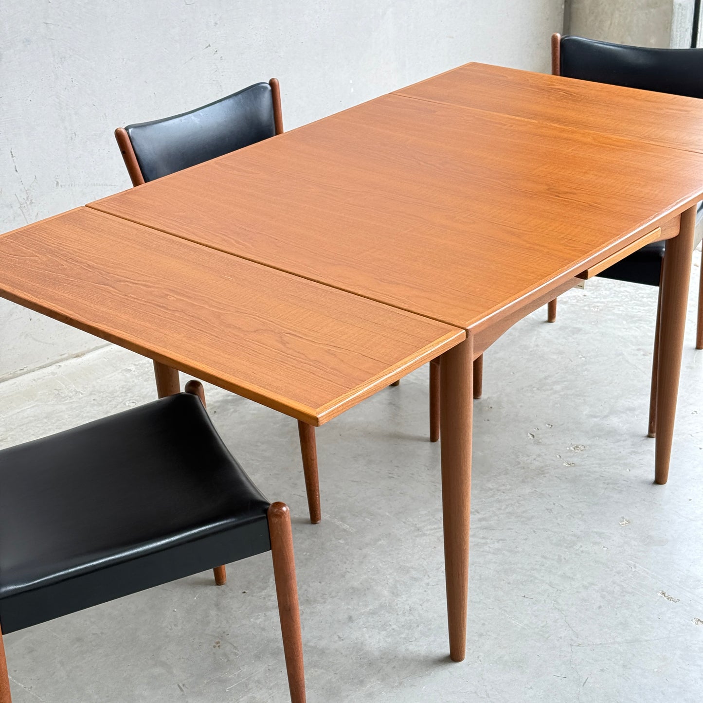 A Fully Restored Parker 140 Square Extension Table (Rediscover) with an extension leaf pulled out is paired with three black leather chairs on a light gray concrete floor against a plain wall.