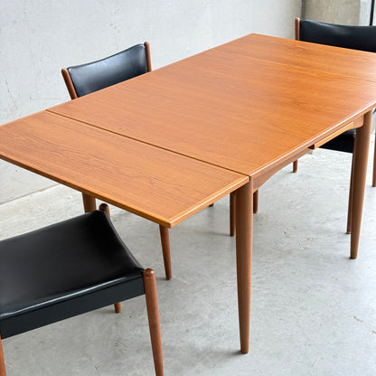 A Fully Restored Parker 140 Square Extension Table (Rediscover) with an extension leaf pulled out is paired with three black leather chairs on a light gray concrete floor against a plain wall.