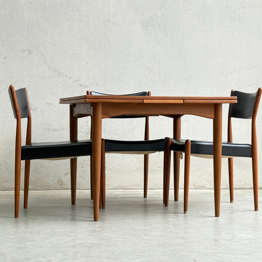 A Fully Restored Parker 140 Square Extension Table (Rediscover) with four black-cushioned chairs sits neatly on a light concrete floor against a plain textured wall.