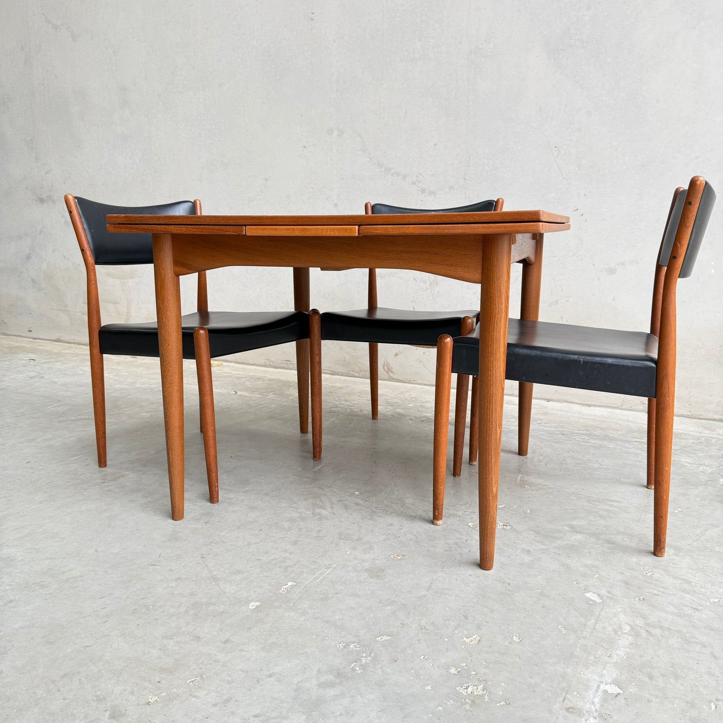 A Fully Restored Parker 140 Square Extension Table (Rediscover) with rounded edges is paired with four matching chairs featuring black seats and backs, set against a plain light gray wall and concrete floor.