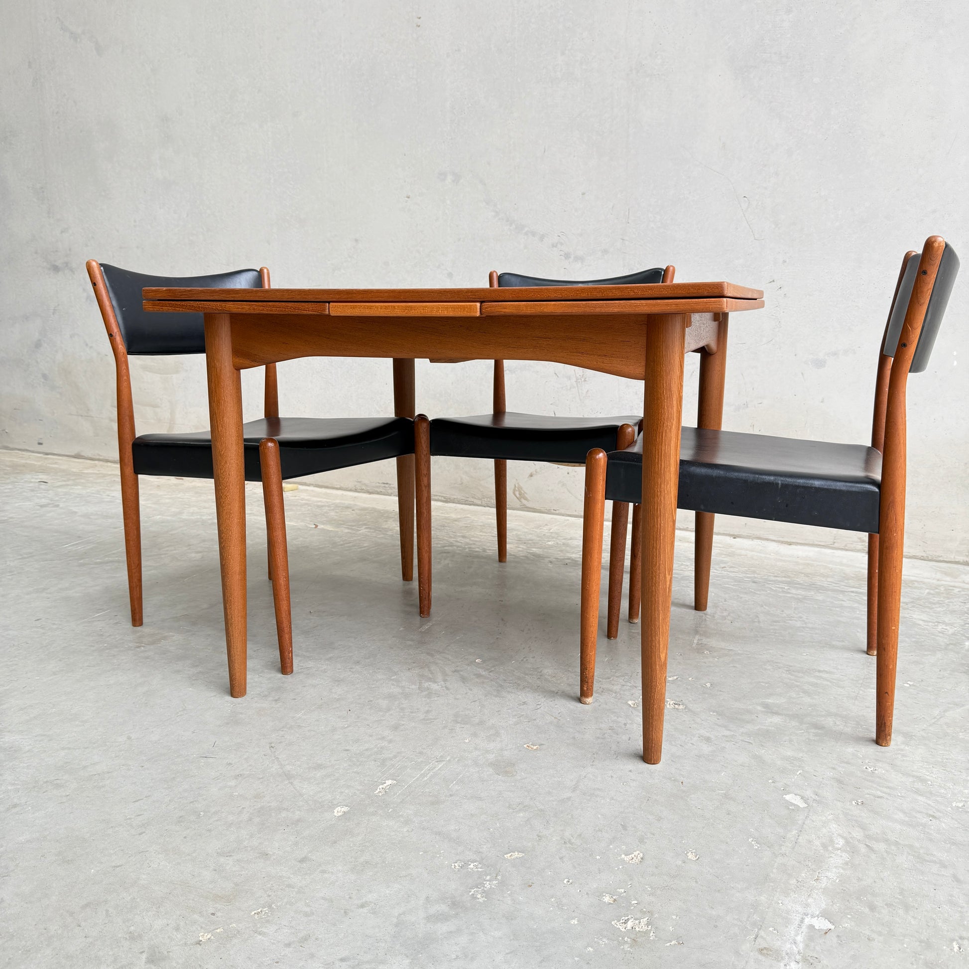 A Fully Restored Parker 140 Square Extension Table (Rediscover) with rounded edges is paired with four matching chairs featuring black seats and backs, set against a plain light gray wall and concrete floor.