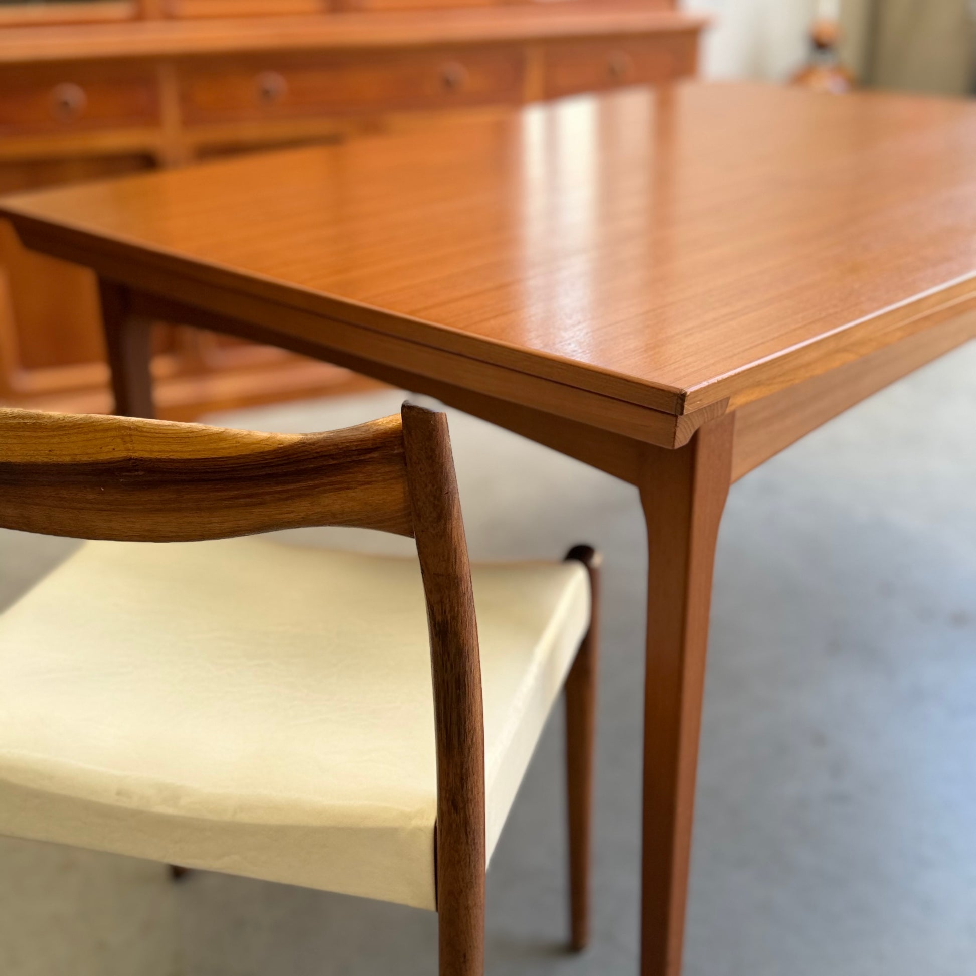 A close-up of the Fler 64 Rectangular extension Table (Refine) with tapered legs, paired with a matching wooden chair featuring a light cushion. A vintage-inspired sideboard appears blurred in the background.