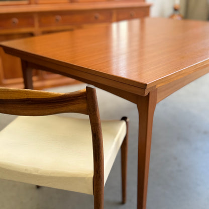 A close-up of the Fler 64 Rectangular extension Table (Refine) with tapered legs, paired with a matching wooden chair featuring a light cushion. A vintage-inspired sideboard appears blurred in the background.