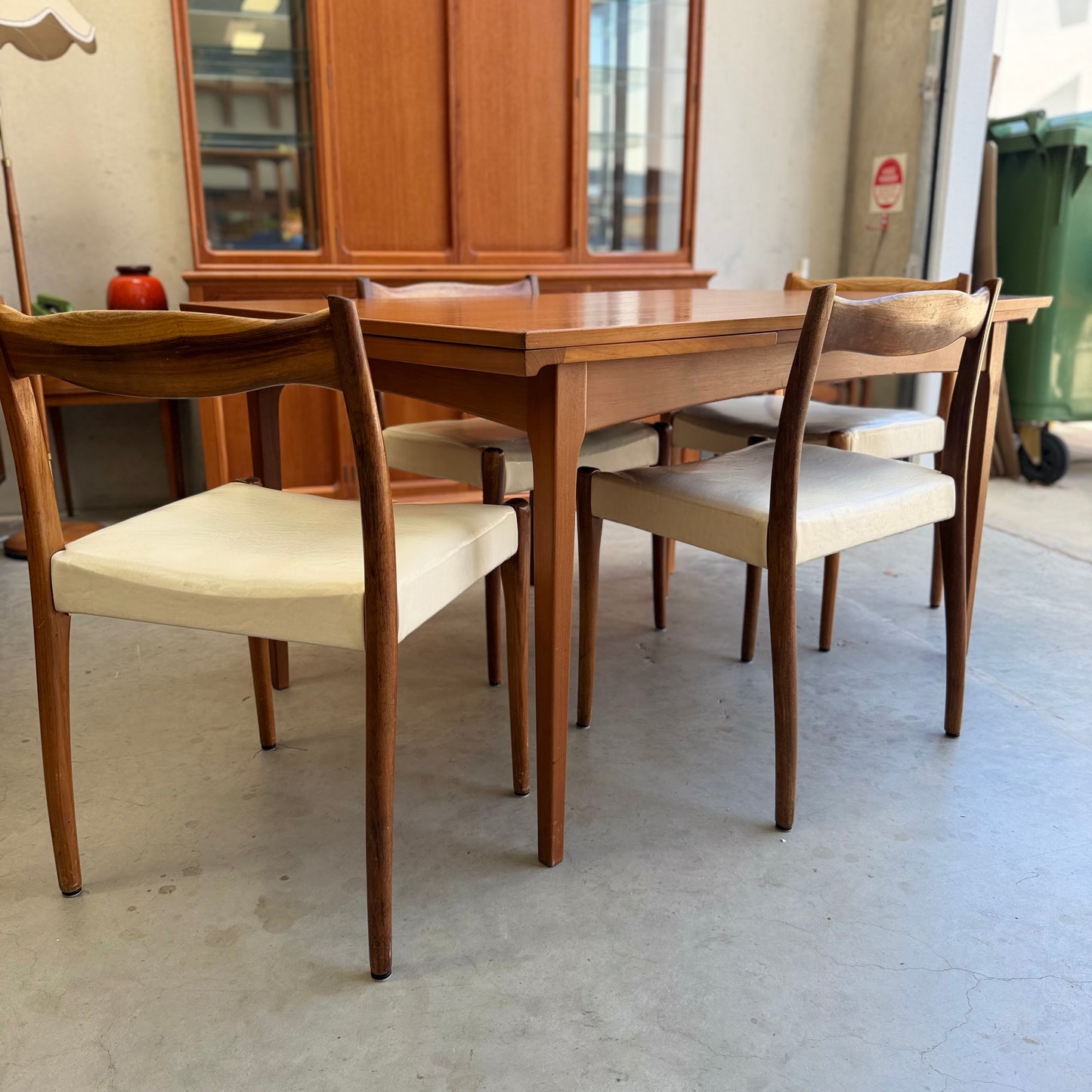 A Fler 64 Rectangular extension Table (Refine) with four matching chairs featuring white cushions is placed in a bright room, alongside a large cabinet, window, and green trash bin in the background.