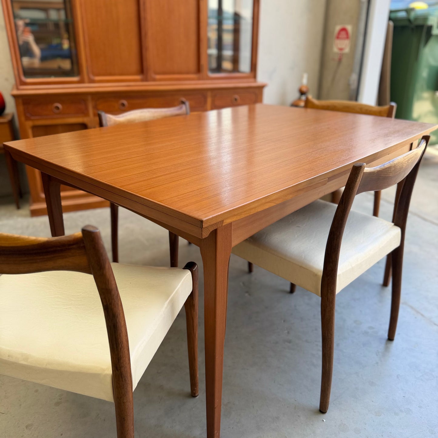 The Fler 64 Rectangular Extension Table (Refine) with three wooden chairs featuring white cushions stands in a room with a wooden cabinet and fire extinguisher, highlighting classic vintage Australian design.
