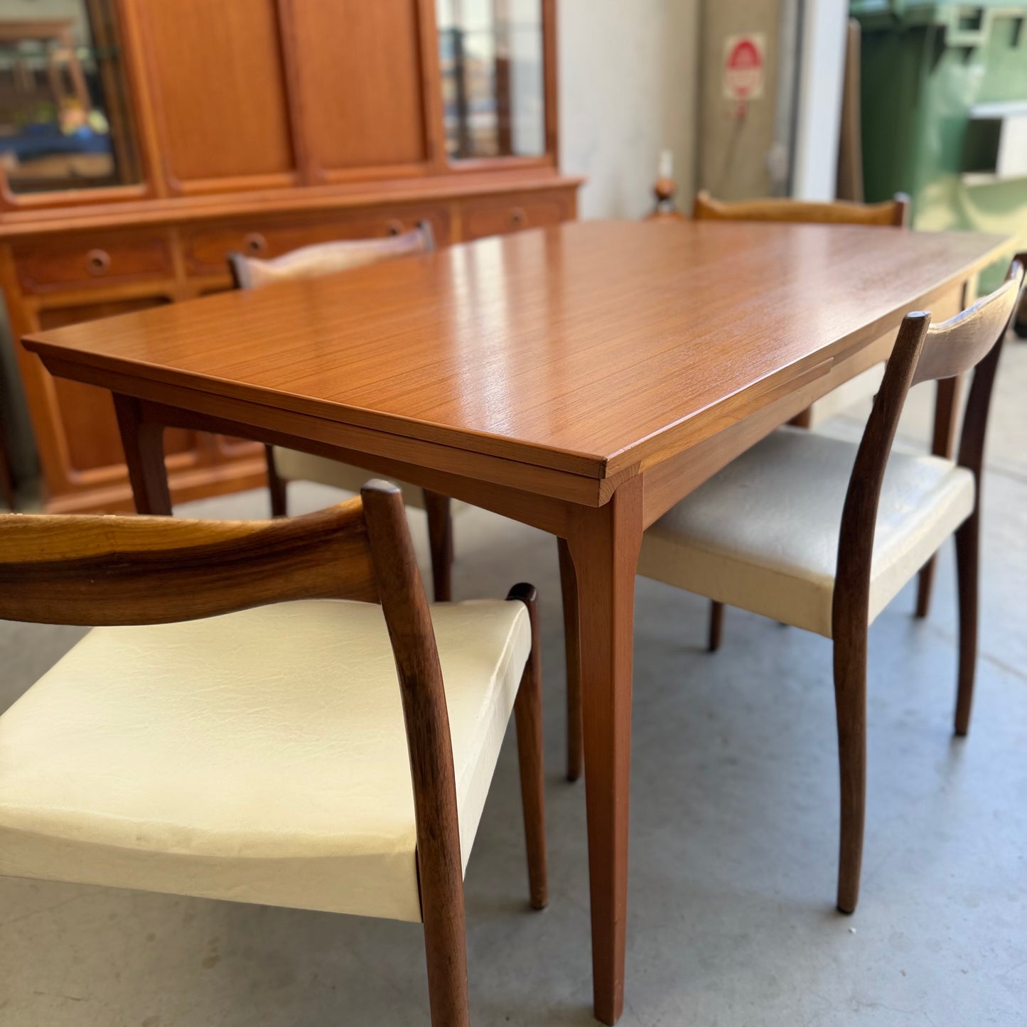 The Fler 64 Rectangular extension Table (Refine) set features four chairs with white seats and wooden backs in a vintage Australian mid-century style, shown in a room with a wooden cabinet and other furniture.
