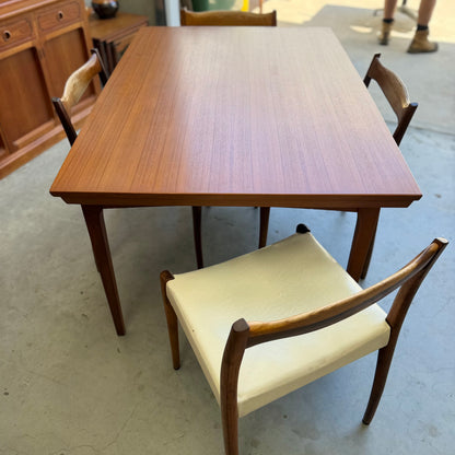 The Fler 64 Rectangular extension Table (Refine) is paired with three wooden chairs—one with a white cushion seat—set on a concrete floor, with more wooden furniture visible in the background.