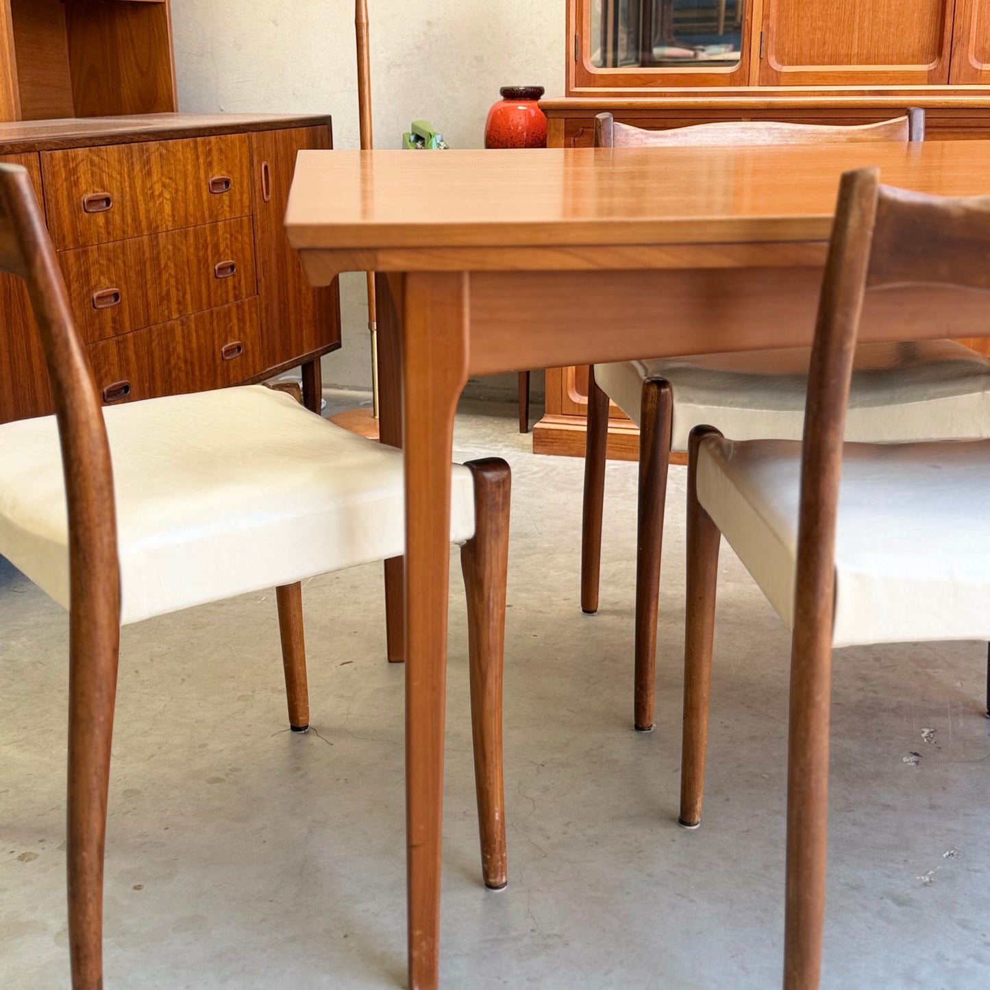 A Fler 64 Rectangular Extension Table (Refine) with matching chairs and white cushions sits on a concrete floor, surrounded by mid-century modern wooden cabinets and a red vase, highlighting vintage Australian design.