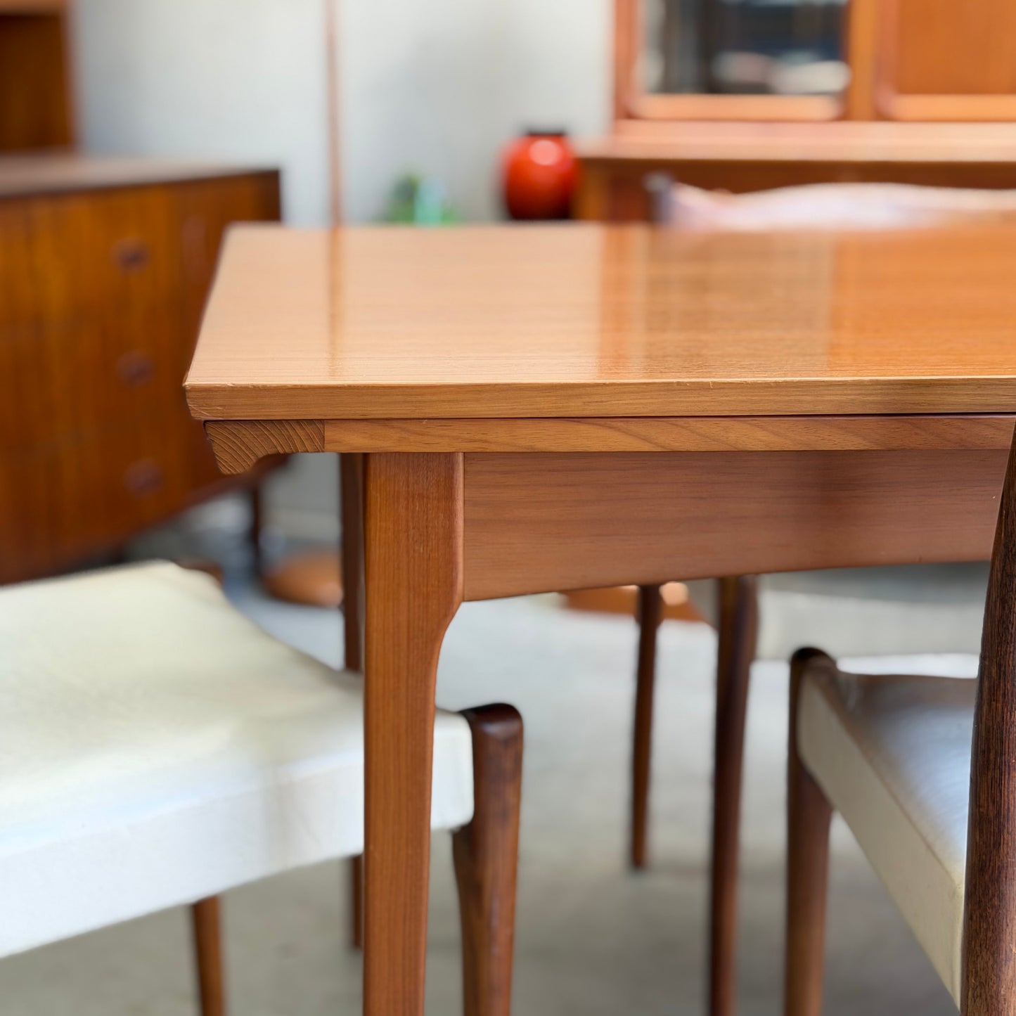 A close-up of the Fler 64 Rectangular Extension Table (Refine) with polished tapered legs, paired with wooden chairs in white and beige. Blurred wooden cabinets and a red vase in the background highlight its vintage Australian design.