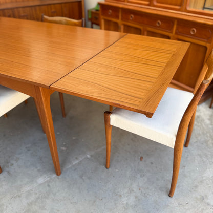 A Fler 64 Rectangular Extension Table (Refine) with a visible leaf and two white-cushioned chairs sits on a concrete floor among other wooden pieces—an elegant touch of vintage Australian style.