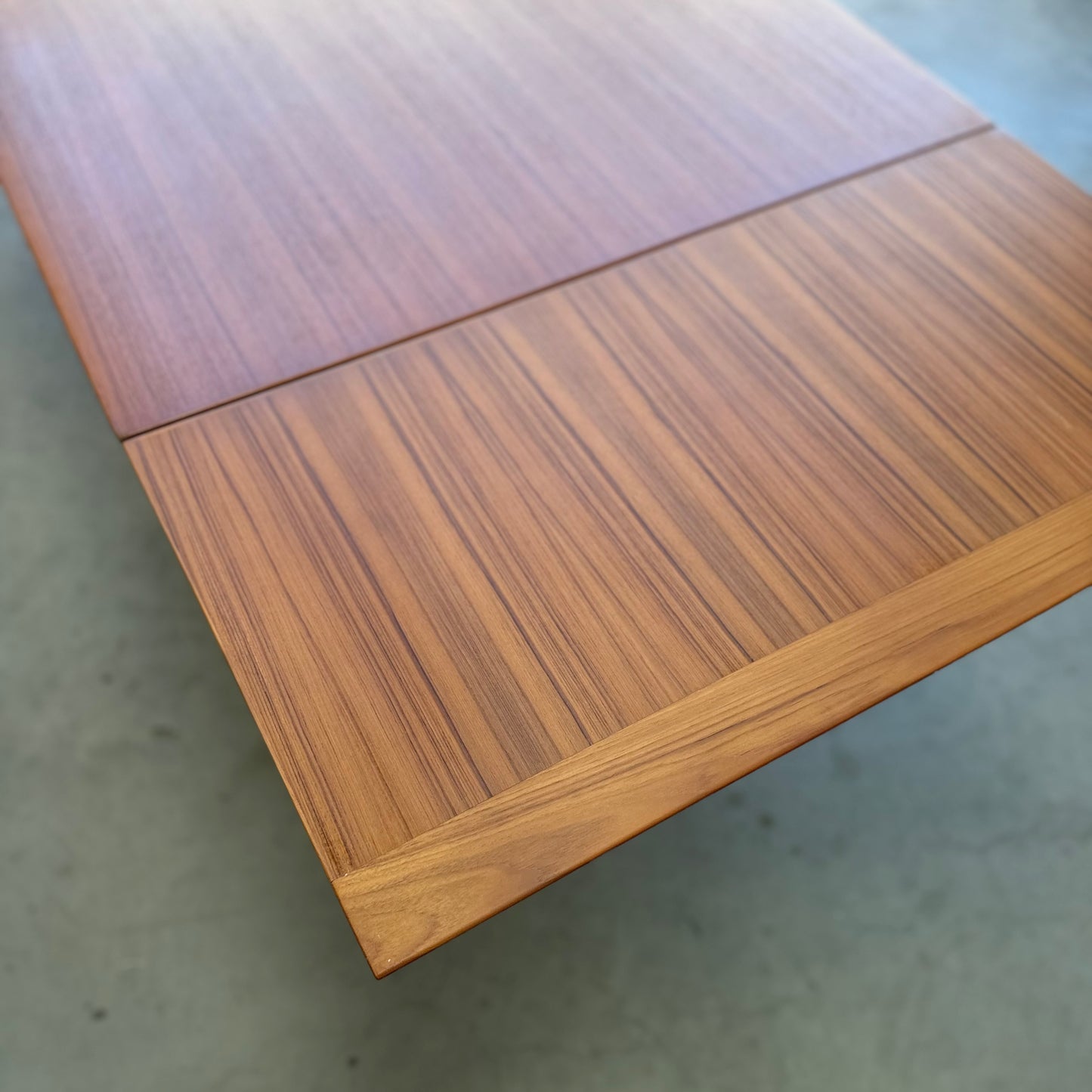 A close-up of the Fler 64 Rectangular extension Table (Refine) highlights its smooth wood surface and visible grain, with a seam across the width showing its extendable mid-century modern design, set on a plain gray floor.