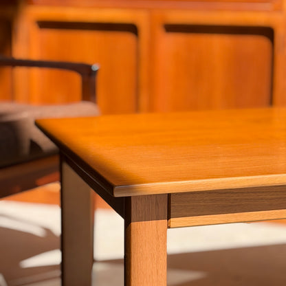 A close-up of the Mid Century Teak Danish Style Coffee Table – Circa 1970s (Revive), sunlight casting shadows on its surface. A wooden chair and wood-paneled wall in the background create a warm, cozy ambiance.