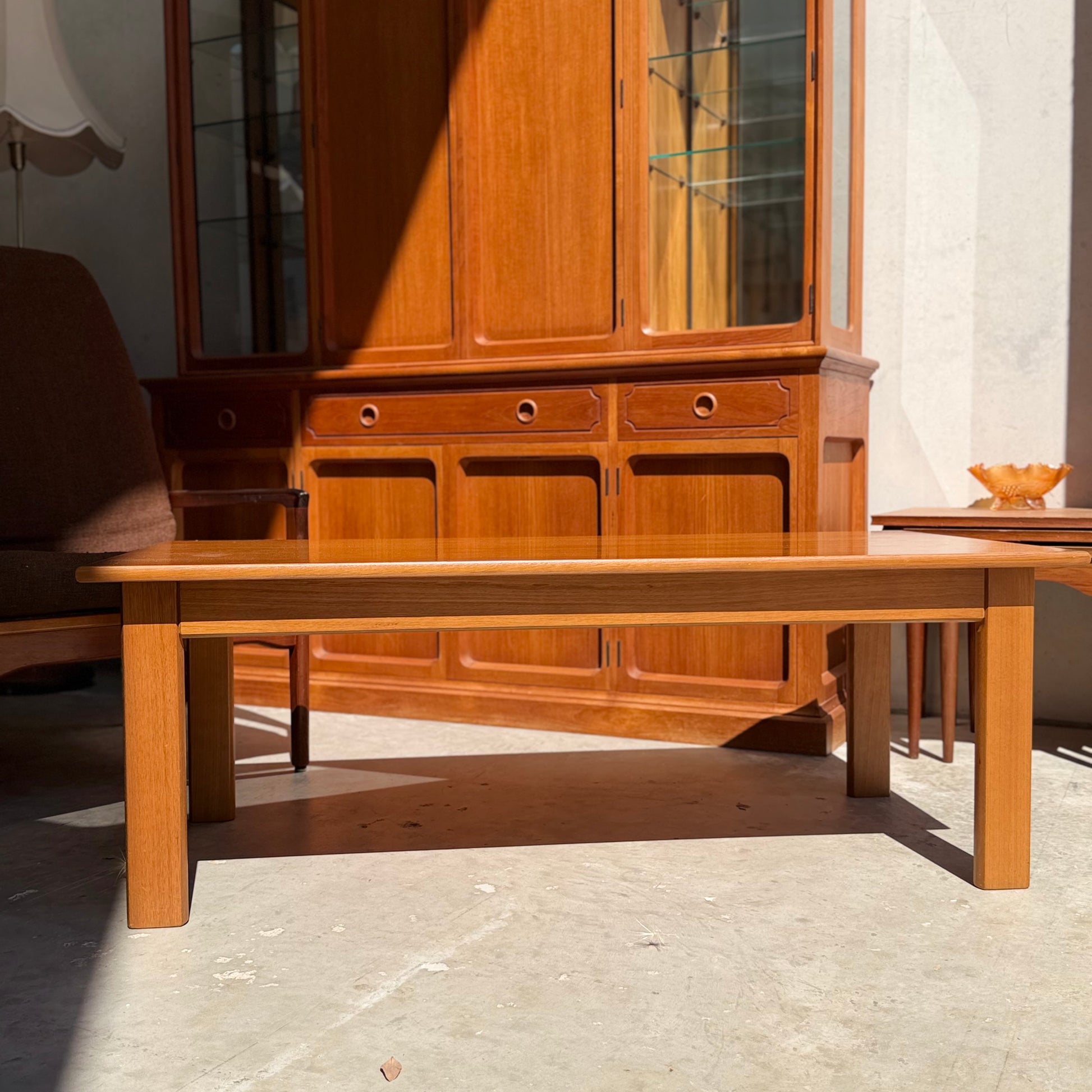 A Mid Century Teak Danish Style Coffee Table – Circa 1970s (Revive) rests in sunlight on a concrete floor, before a large glass-doored cabinet. Nearby, a brown armchair and side table with an orange bowl complete this vintage Scandinavian scene.