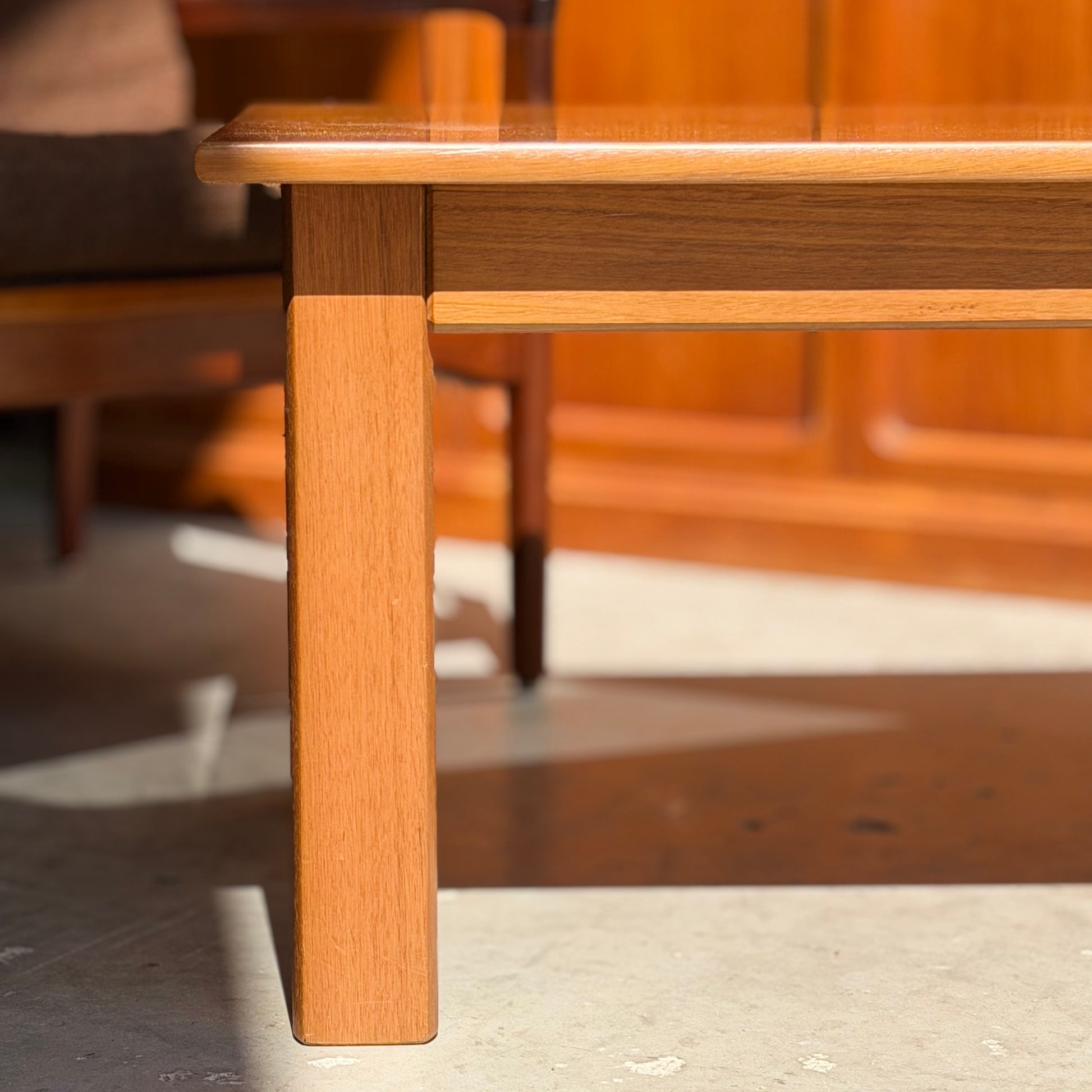 A close-up of the Mid Century Teak Danish Style Coffee Table – Circa 1970s (Revive) shows its leg and edge in sunlight on a smooth floor, with a wooden chair and paneled wall evoking vintage Scandinavian design in the background.