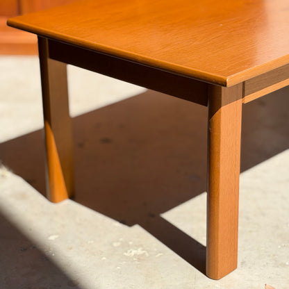 The Mid Century Teak Danish Style Coffee Table – Circa 1970s (Revive) features square legs and a smooth finish, casting a bold shadow as it rests on a sunlit concrete floor.