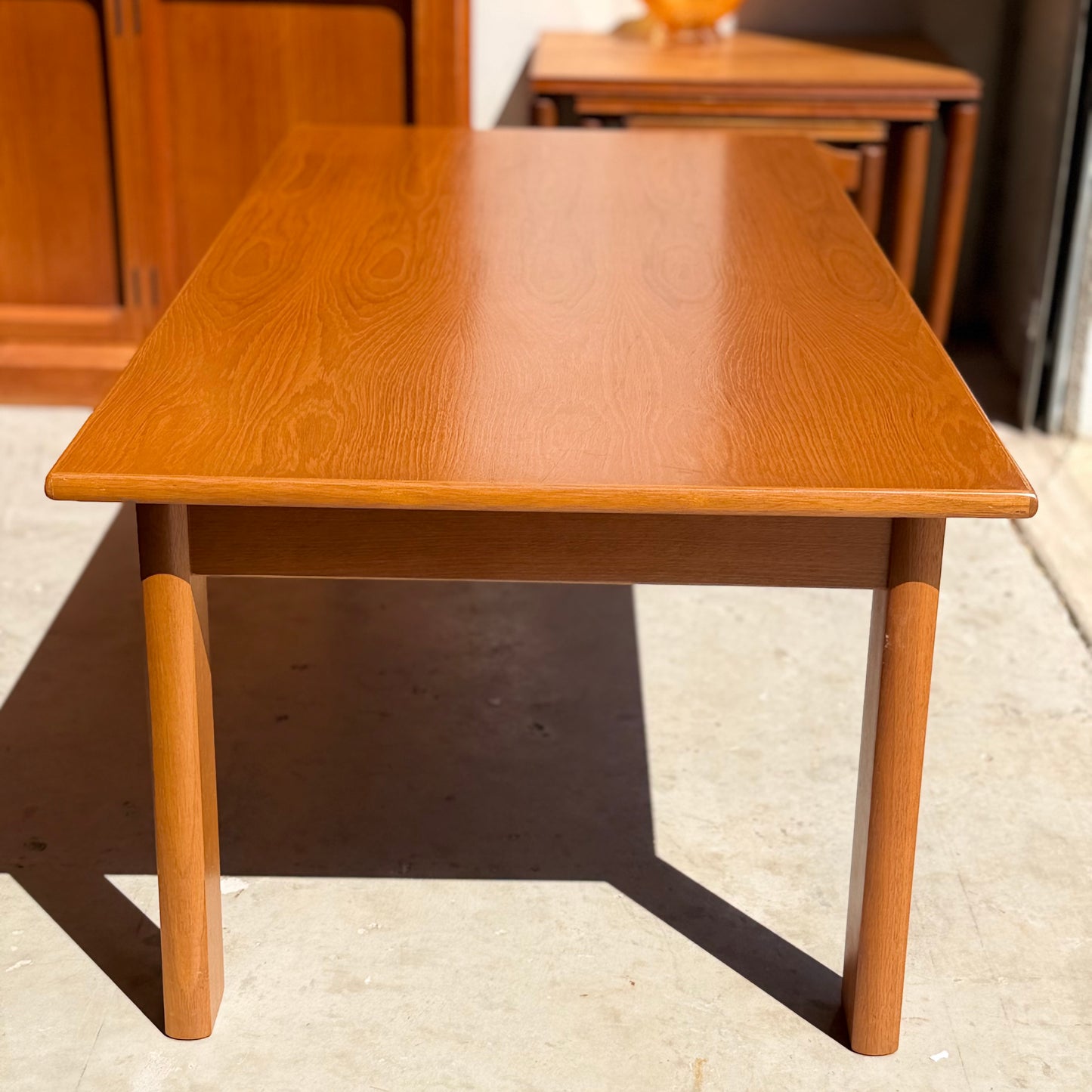 A Mid Century Teak Danish Style Coffee Table (circa 1970s, Revive) with a smooth, light brown finish and four sturdy legs sits on a concrete floor in a sunlit space, surrounded by other vintage Scandinavian furniture.