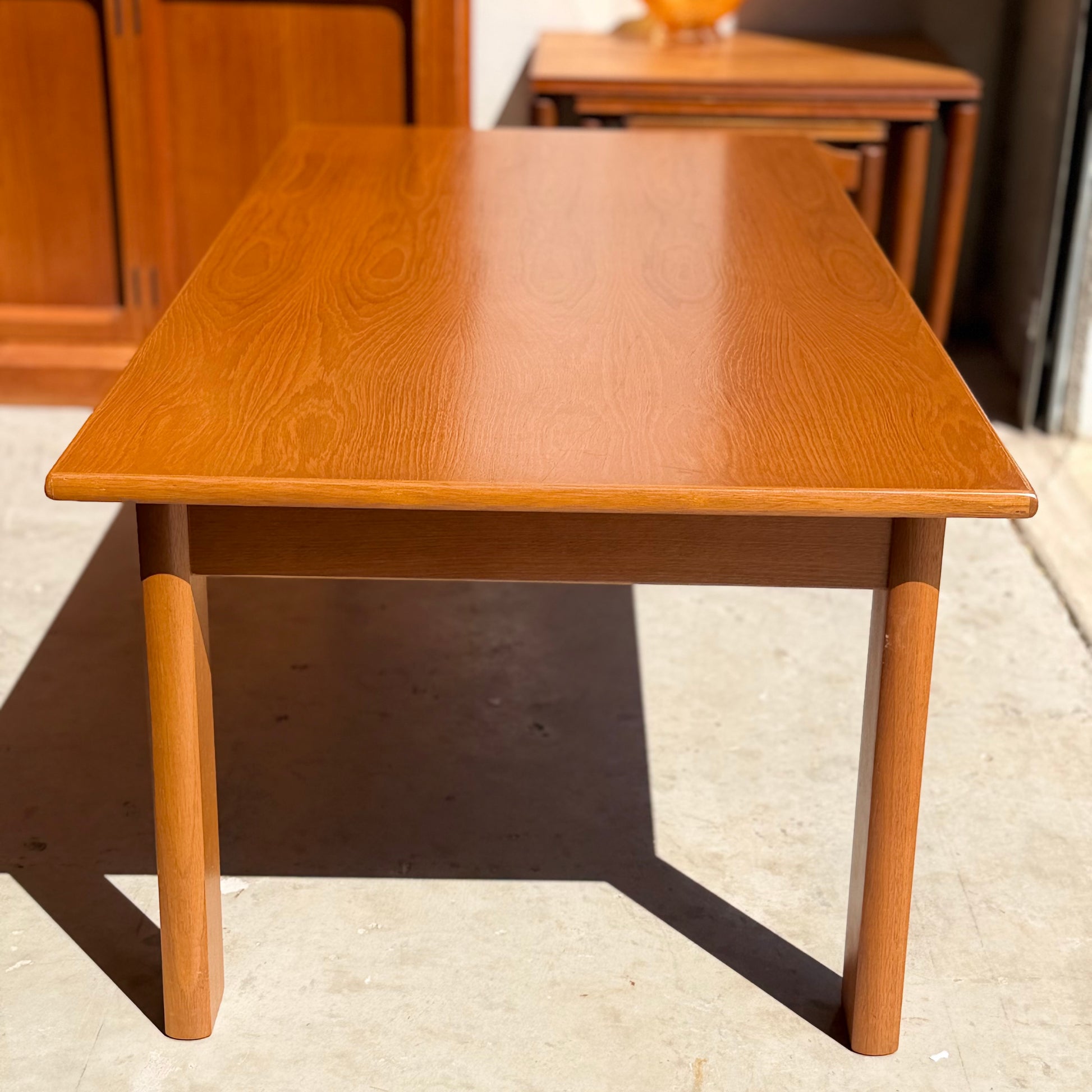 A Mid Century Teak Danish Style Coffee Table (circa 1970s, Revive) with a smooth, light brown finish and four sturdy legs sits on a concrete floor in a sunlit space, surrounded by other vintage Scandinavian furniture.