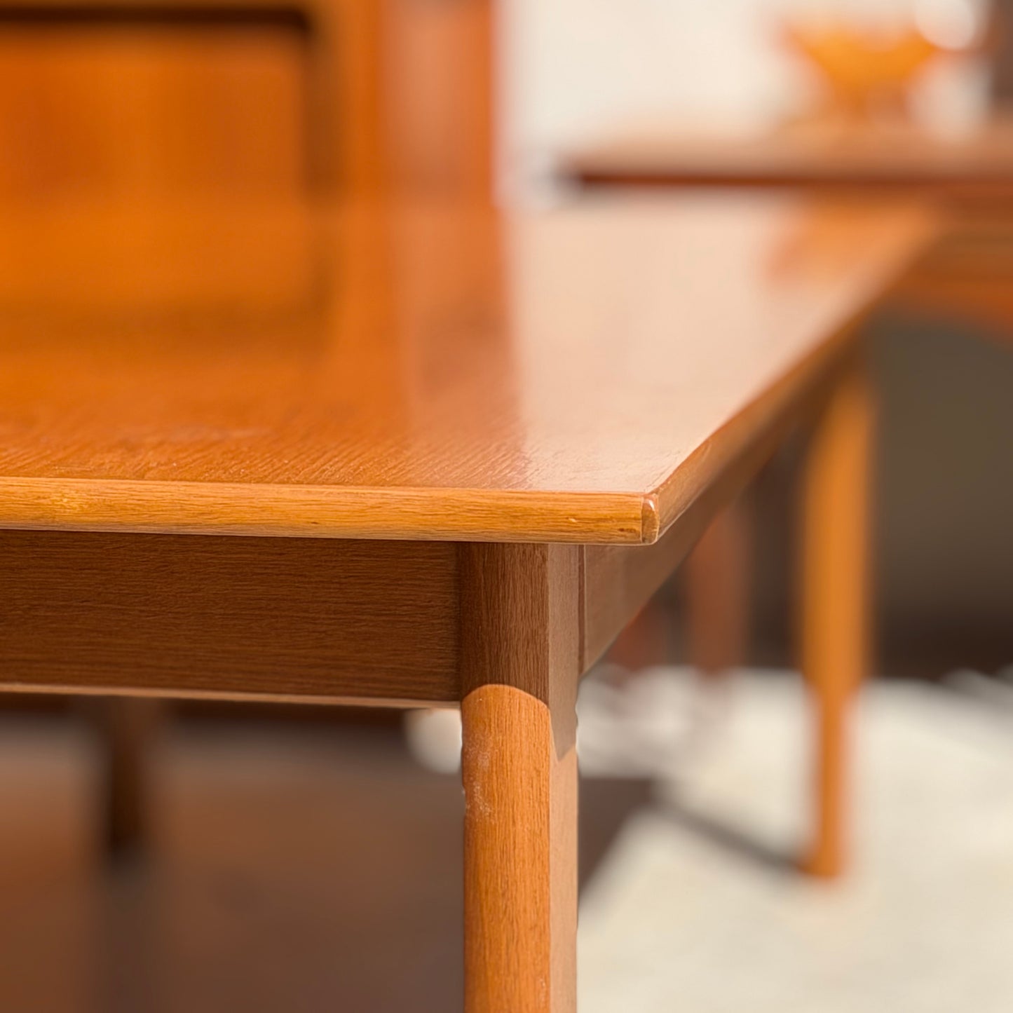 Close-up of the Mid Century Teak Danish Style Coffee Table – Circa 1970s (Revive) with smooth, rounded edges and a polished finish. Sunlight highlights its rich wood grain, while matching vintage Scandinavian furniture is blurred in the background.