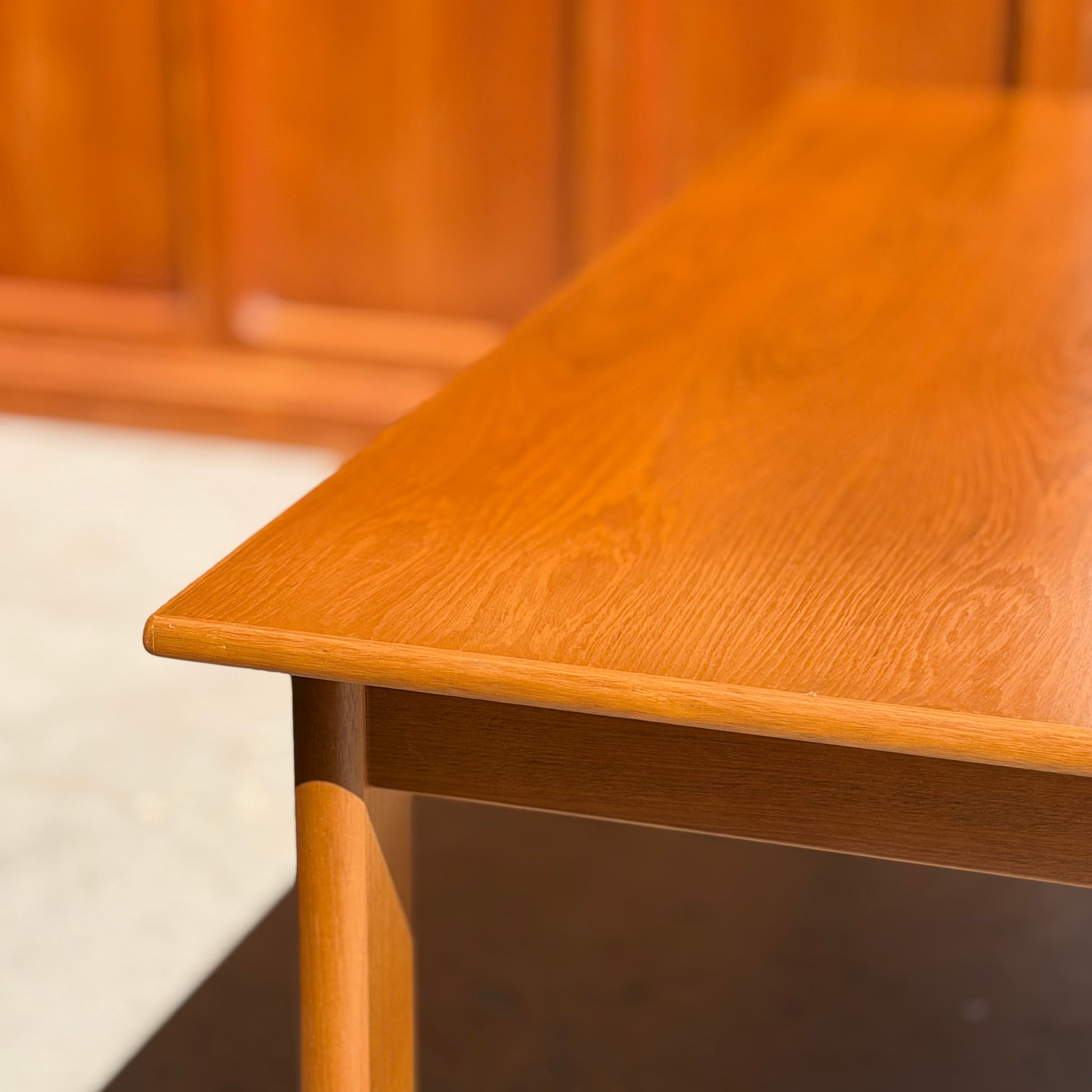 A close-up of the Mid Century Teak Danish Style Coffee Table – Circa 1970s (Revive) shows its smooth, light brown finish and sunlit wood grain, with a matching Danish-style wooden wall softly blurred in the background.
